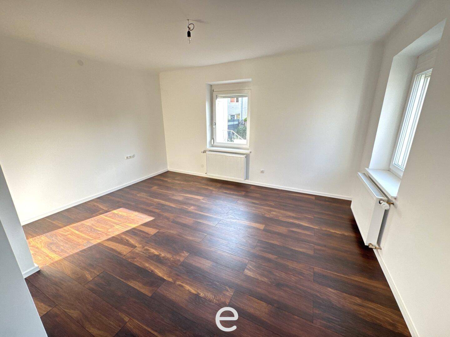 Helles Zimmer mit dunklem Laminatboden und zwei Fenstern, die für eine gute Belichtung sorgen.