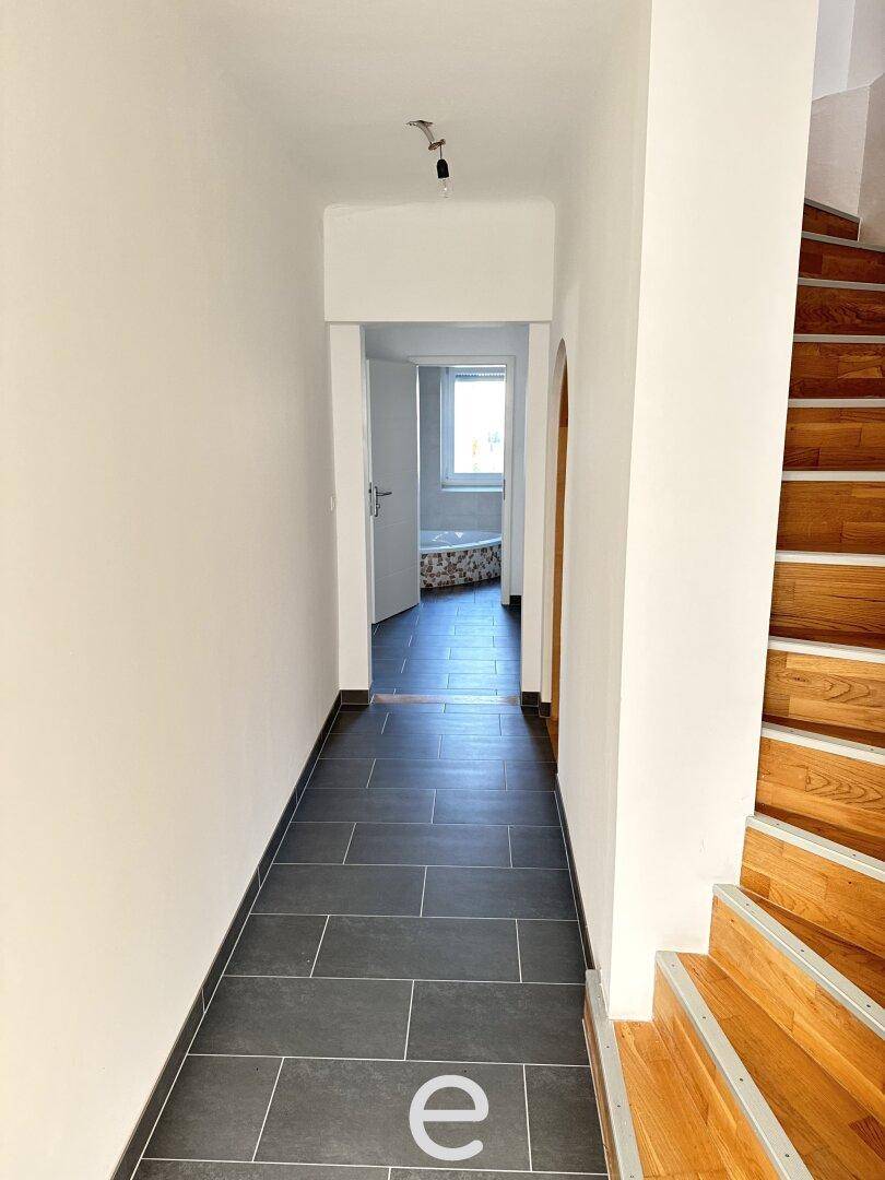 Langer Flur mit dunklem Fliesenboden und einer Holztreppe, die in die oberen Stockwerke führt.