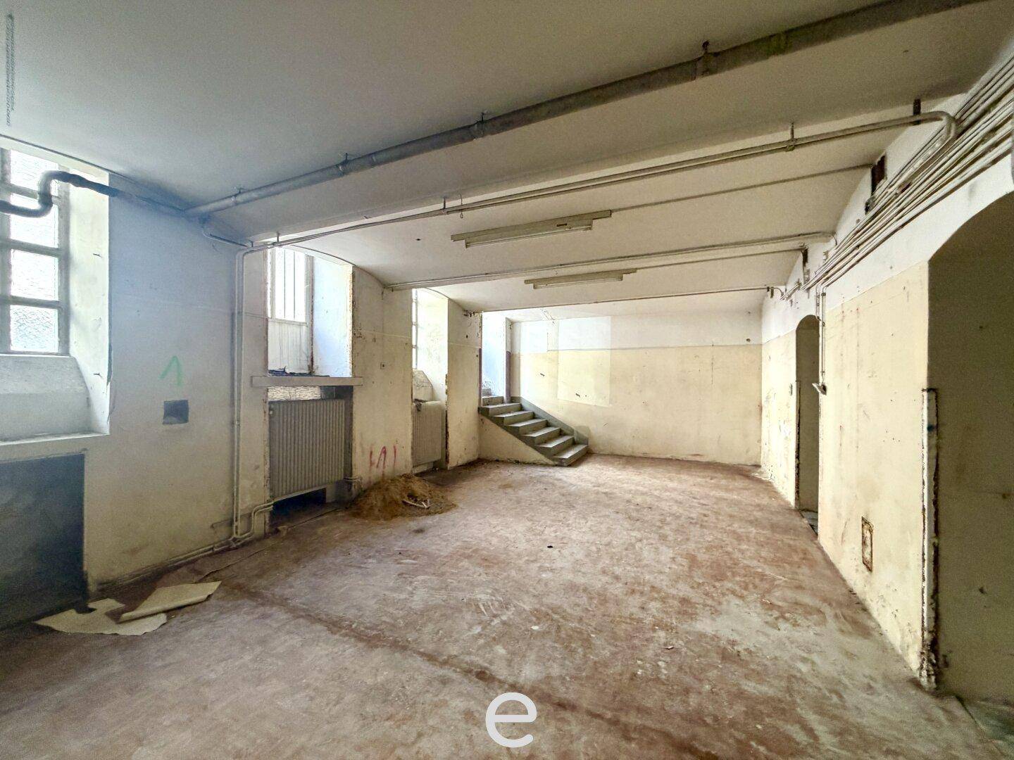 Großer, leerstehender Gewerberaum mit sichtbaren Rohren, Heizkörper und einer Treppe im Hintergrund.