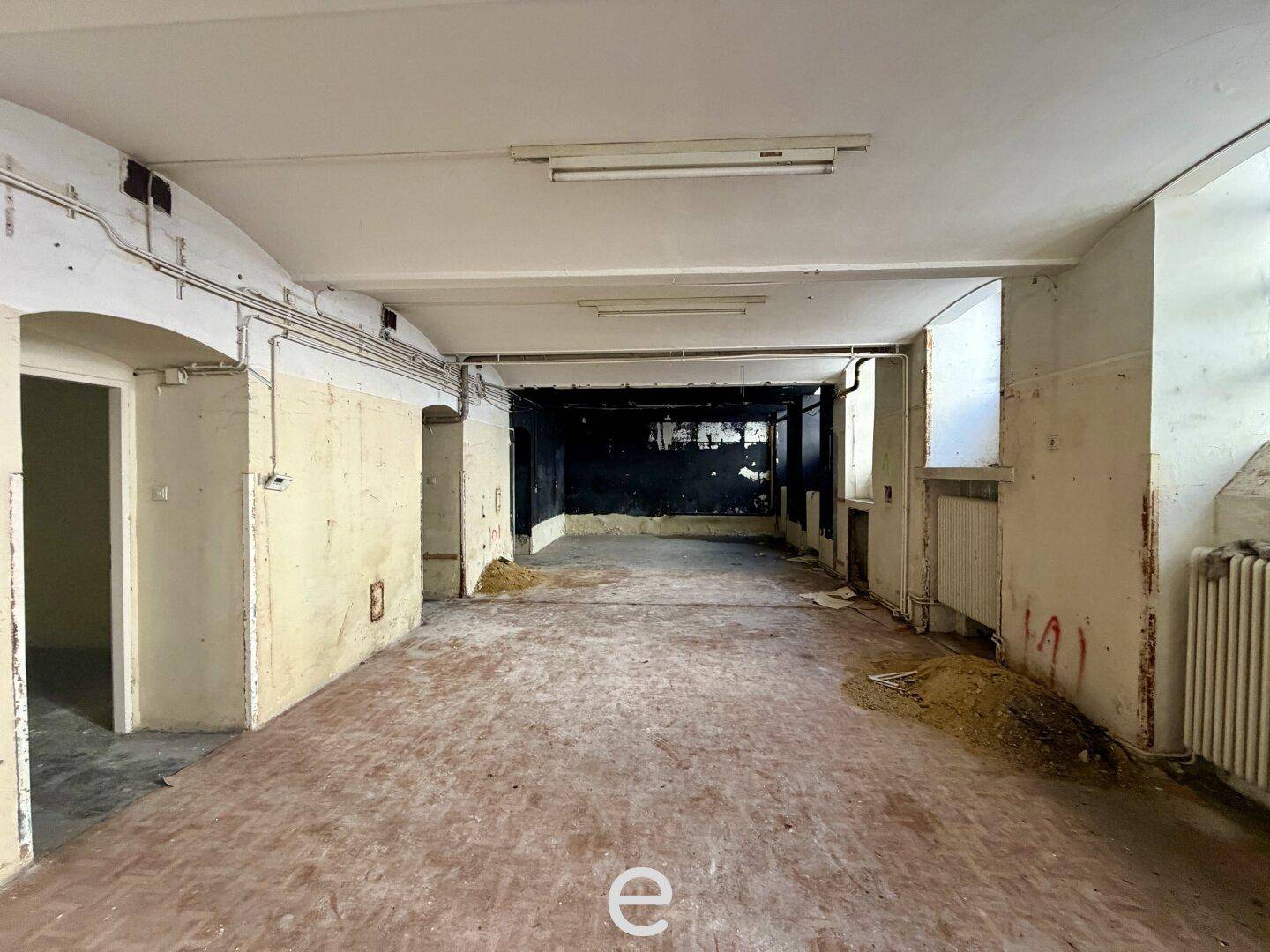 Weitläufiger, unrenovierter Gewerberaum mit dunkler Rückwand und sichtbaren Installationen an der Decke.
