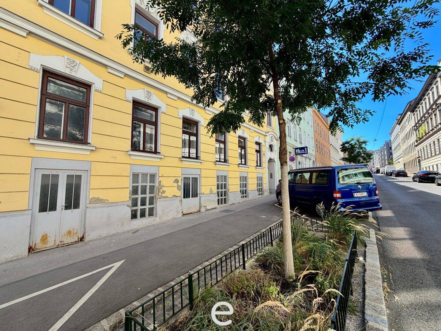 Straßenansicht eines gelben Gebäudes mit Erdgeschosszugang, geparktem Lieferwagen und einem Baum am Straßenrand.
