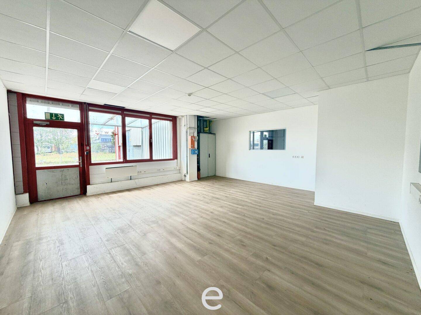 Leerer Büroraum mit Laminatboden, großen Fenstern und Zugang nach draußen.