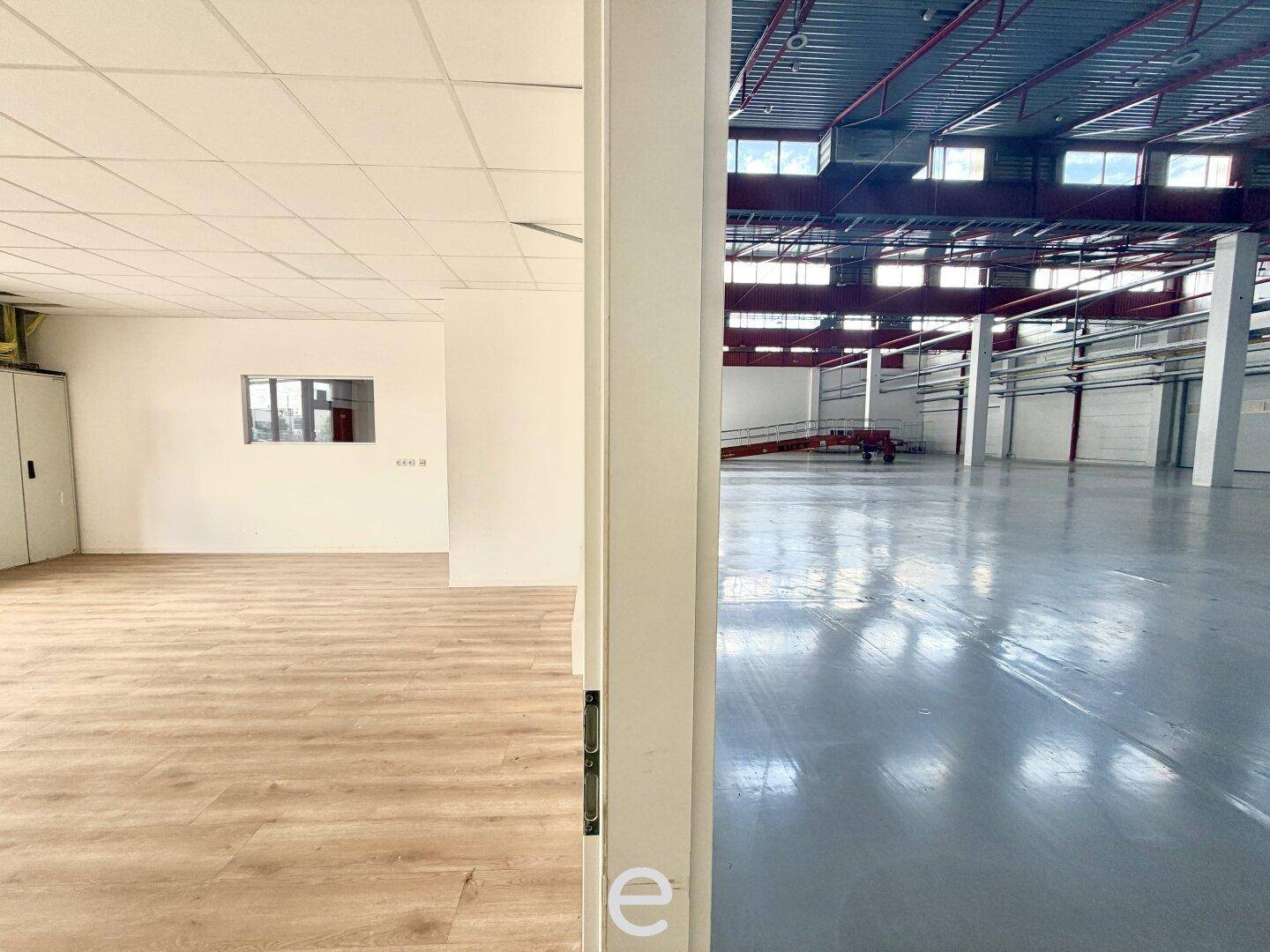 Blick aus einem Büroraum mit Laminatboden in die angrenzende, geräumige Lagerhalle.