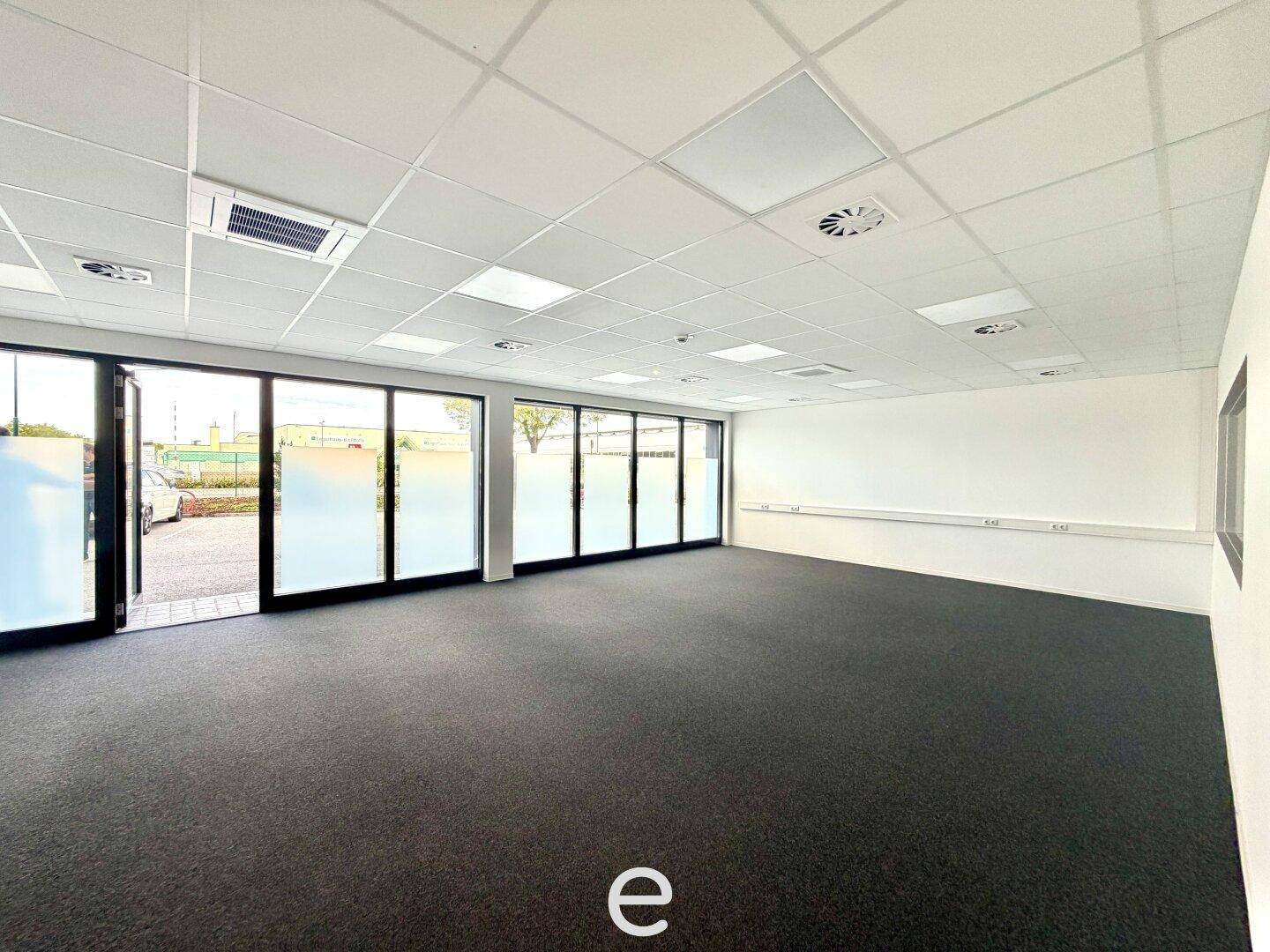 Geräumiger, leerer Büroraum mit dunklem Teppichboden, abgehängter Decke und großen Fenstern.