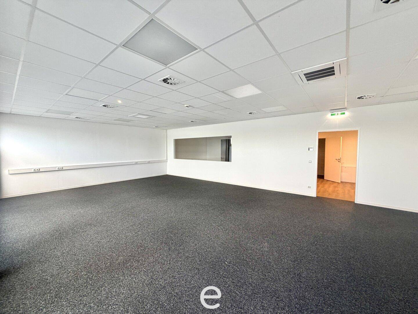 Heller Büroraum mit Teppichboden, abgehängter Decke und einer internen Fensteröffnung zu einem Nebenraum.