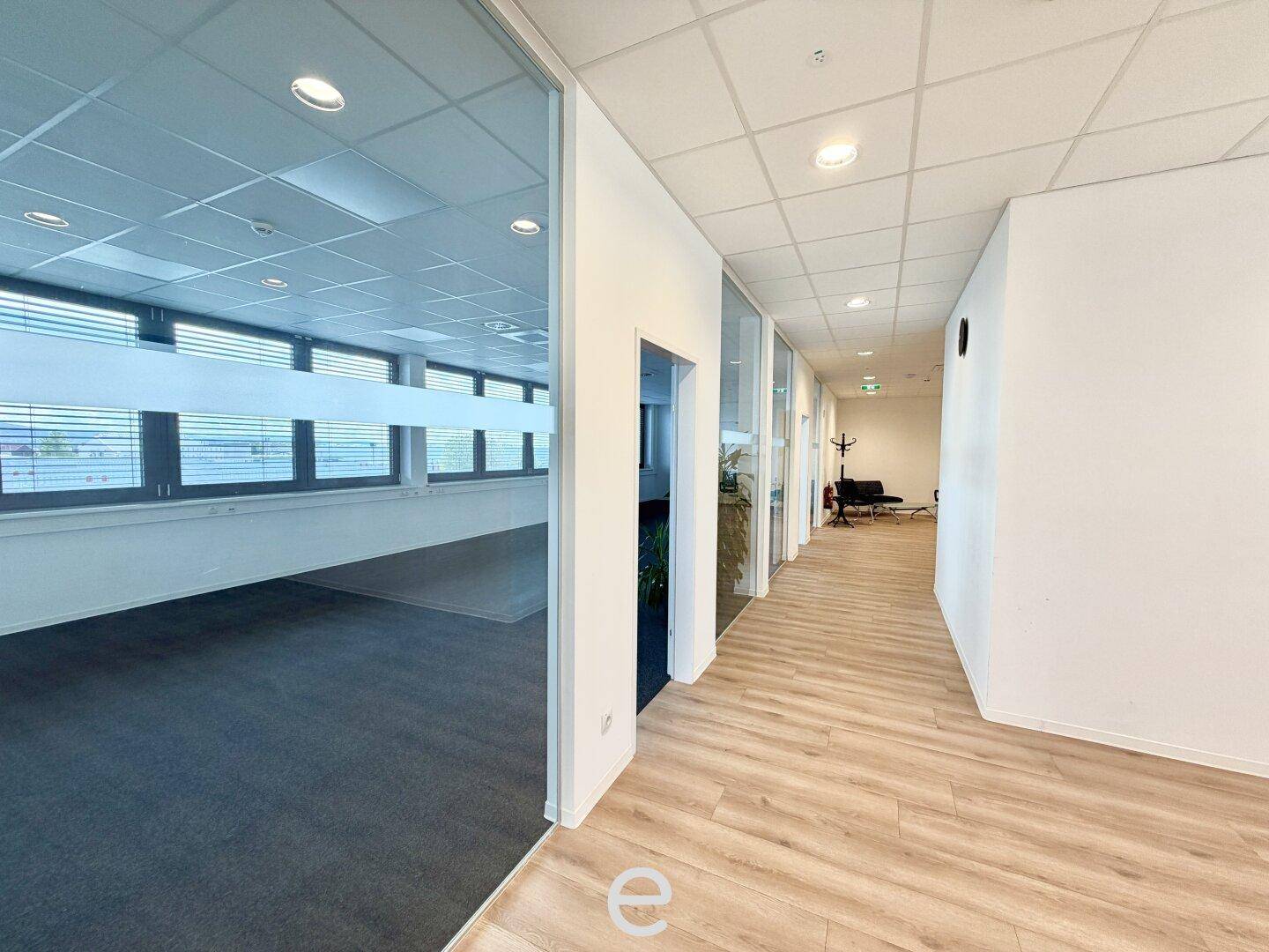Heller Bürogang mit Holzboden und Blick in ein angrenzendes Büro mit großen Fenstern.