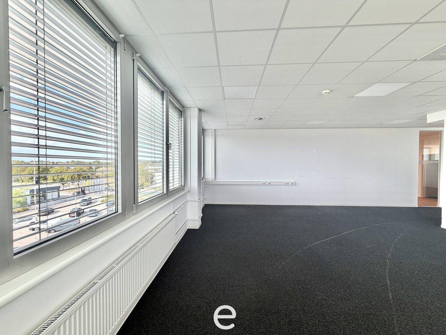 Leeres Büro mit dunklem Teppichboden und einer langen Fensterfront mit Jalousien, die viel Tageslicht hereinlässt.