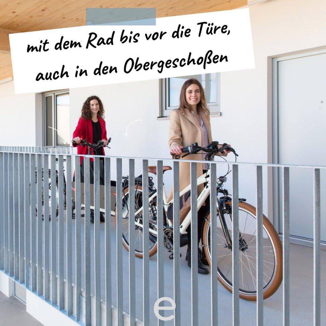 Bewohnerinnen mit Fahrrädern in einem breiten Flur oder auf einem Laubengang des Gebäudes.
