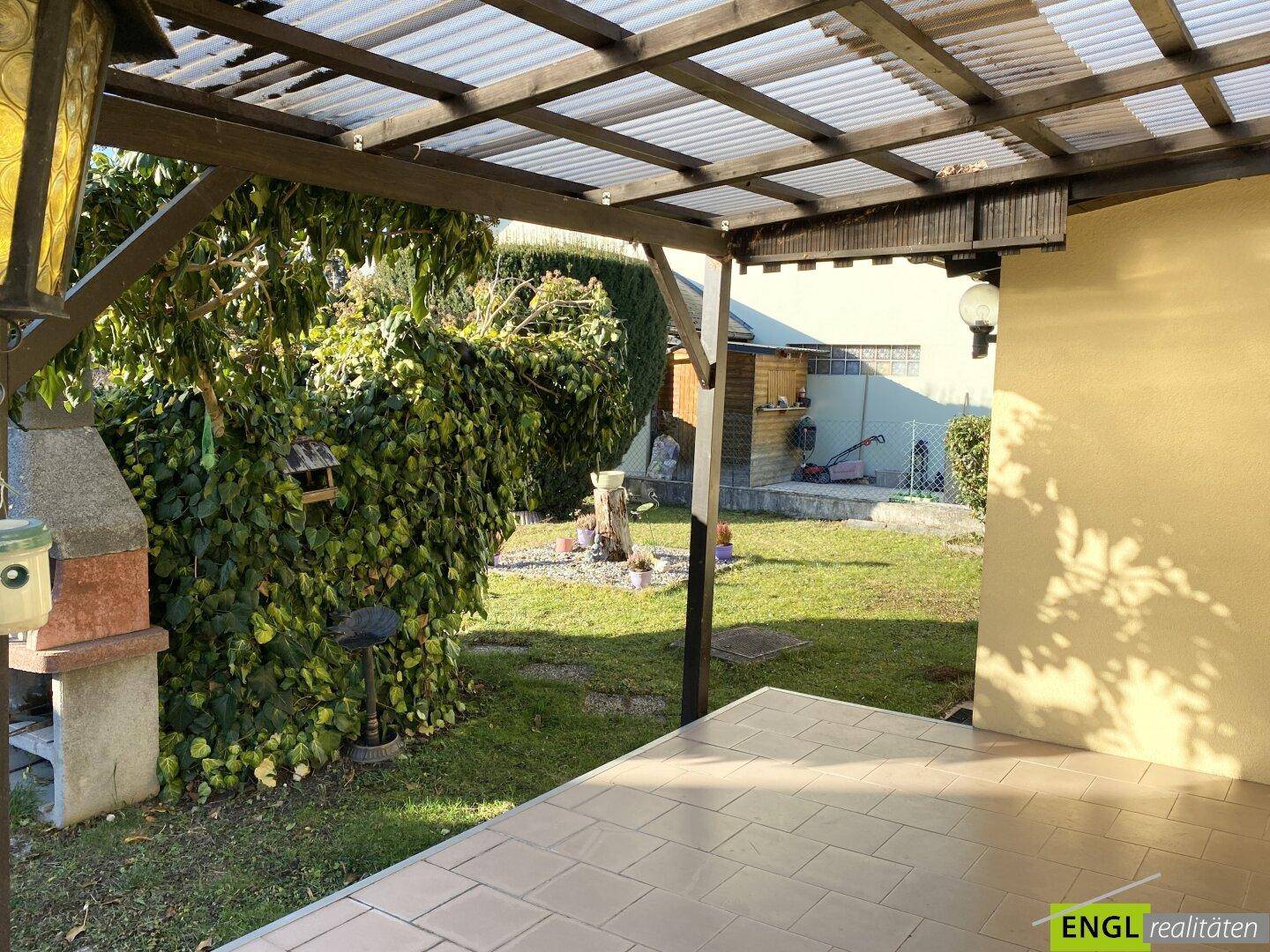 Überdachte Terrasse mit Fliesenboden und integriertem Grillbereich, umgeben von Grün.
