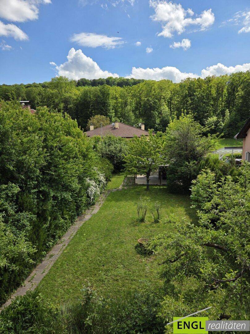 Panoramablick über einen weitläufigen Garten und die umliegende bewaldete Landschaft unter blauem Himmel.