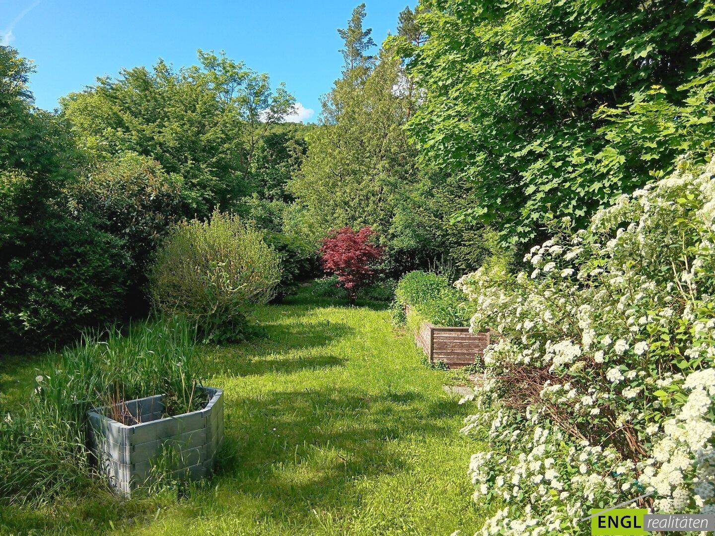 Weitläufige Gartenanlage mit Rasenflächen, Hochbeeten und blühenden Sträuchern, umgeben von Bäumen.