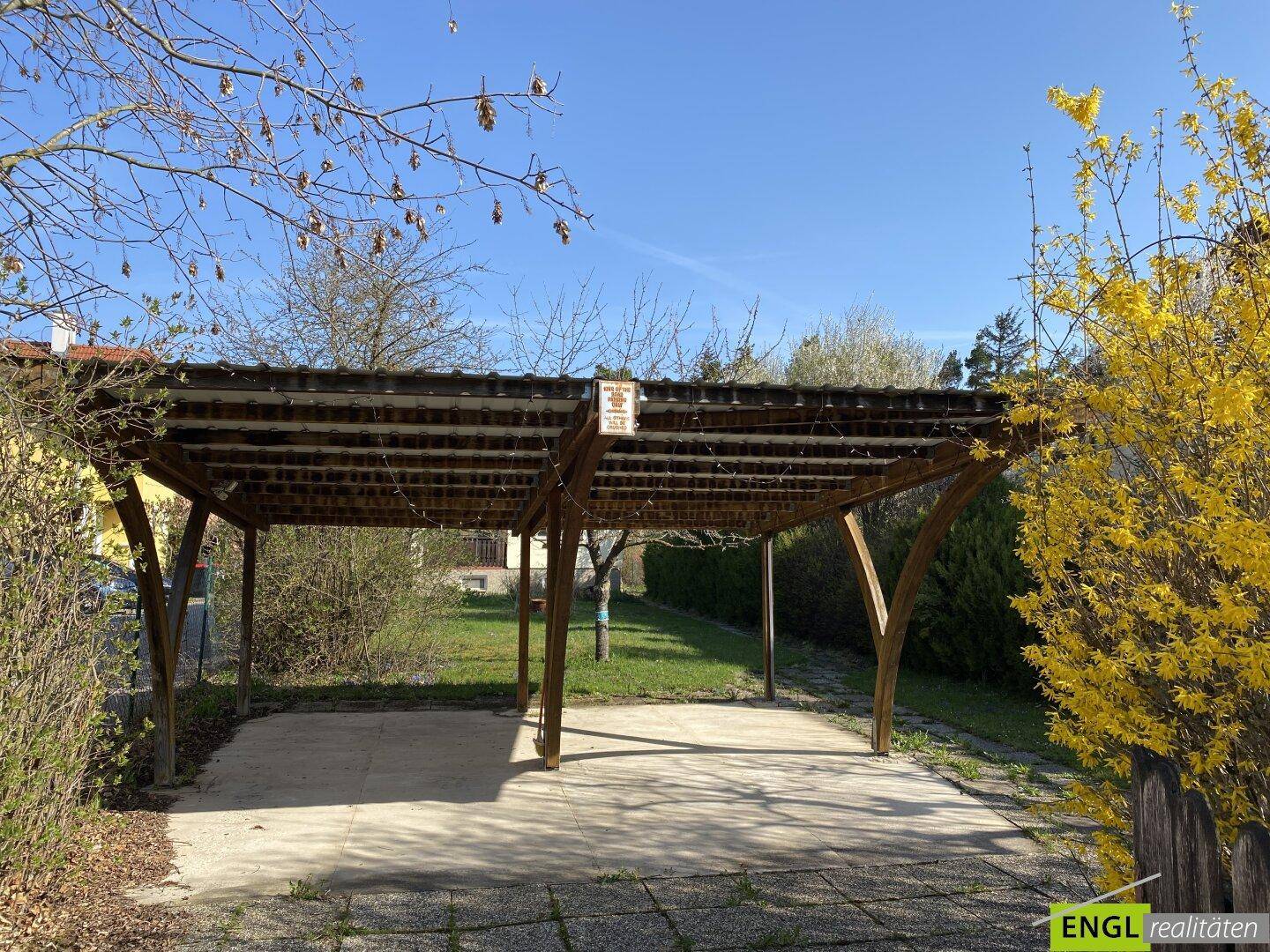 Ein einfacher Holzcarport mit gepflastertem Boden, umgeben von Sträuchern und Bäumen.