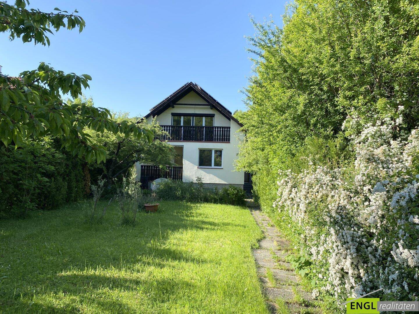 Hausfassade mit Balkon und einem gepflegten Gartenbereich, der von einer Hecke begrenzt wird.