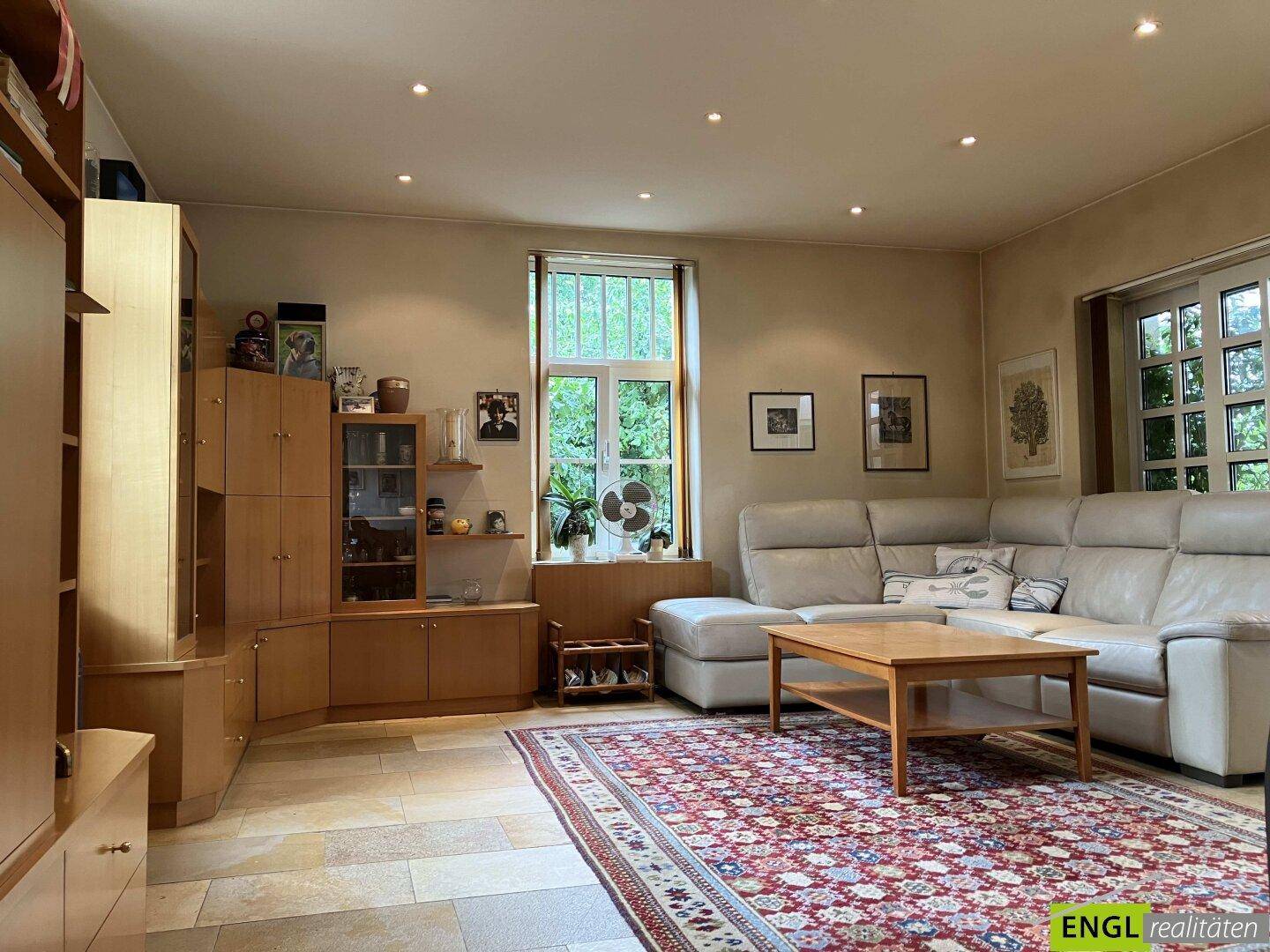 Gemütliches Wohnzimmer mit beigem Sofa, Holzmöbeln und Fliesenboden, hell durch Fenster.