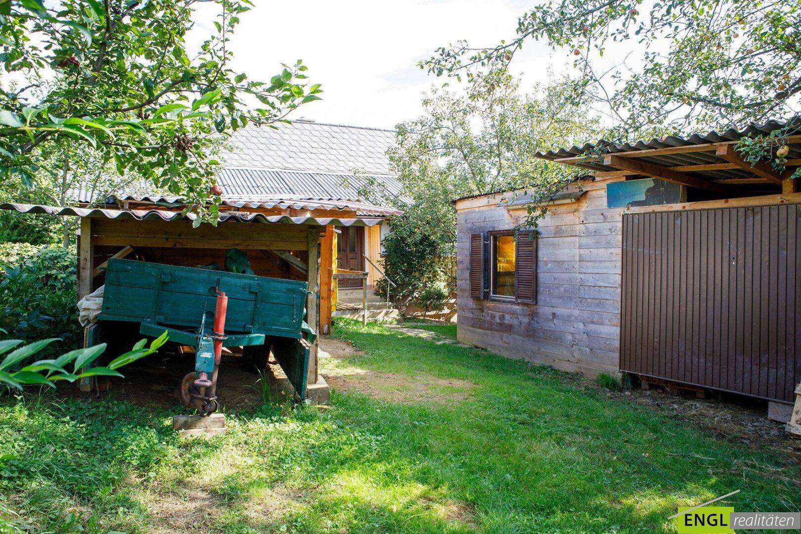 Grüner Gartenbereich mit mehreren Holzschuppen und einem Anhänger, umgeben von Bäumen und Sträuchern.