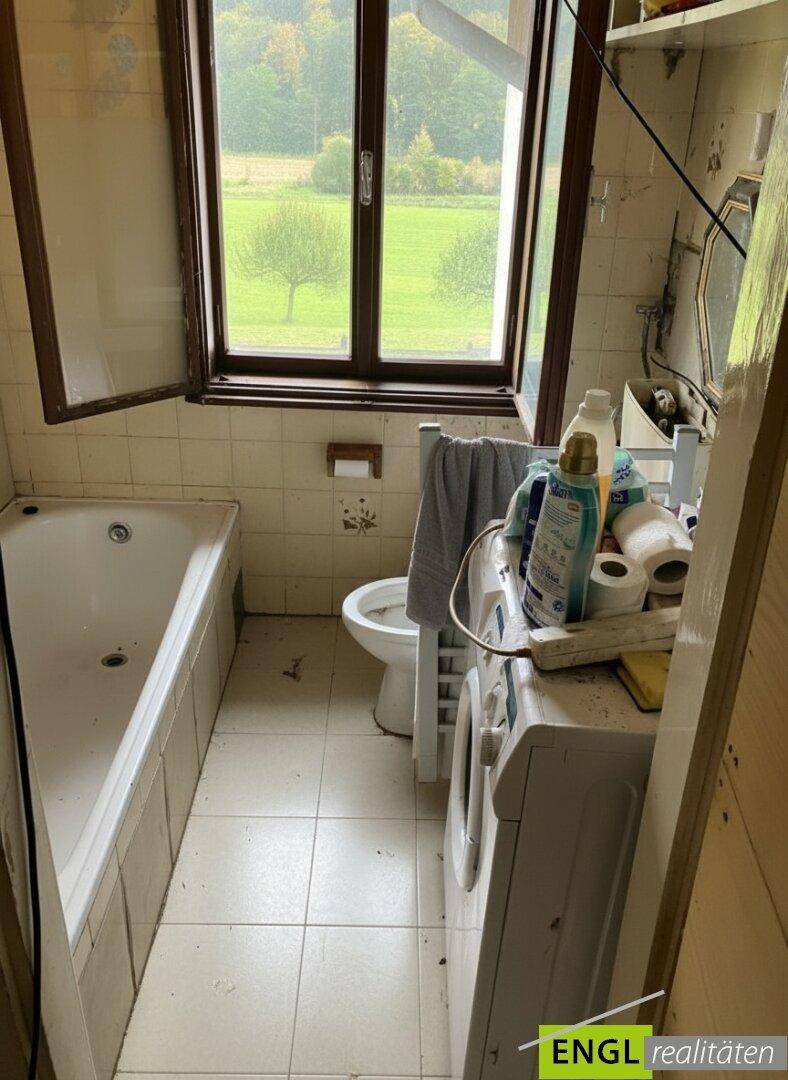 Badezimmer mit Badewanne, Toilette, Waschmaschine und Fenster mit Blick ins Grüne.