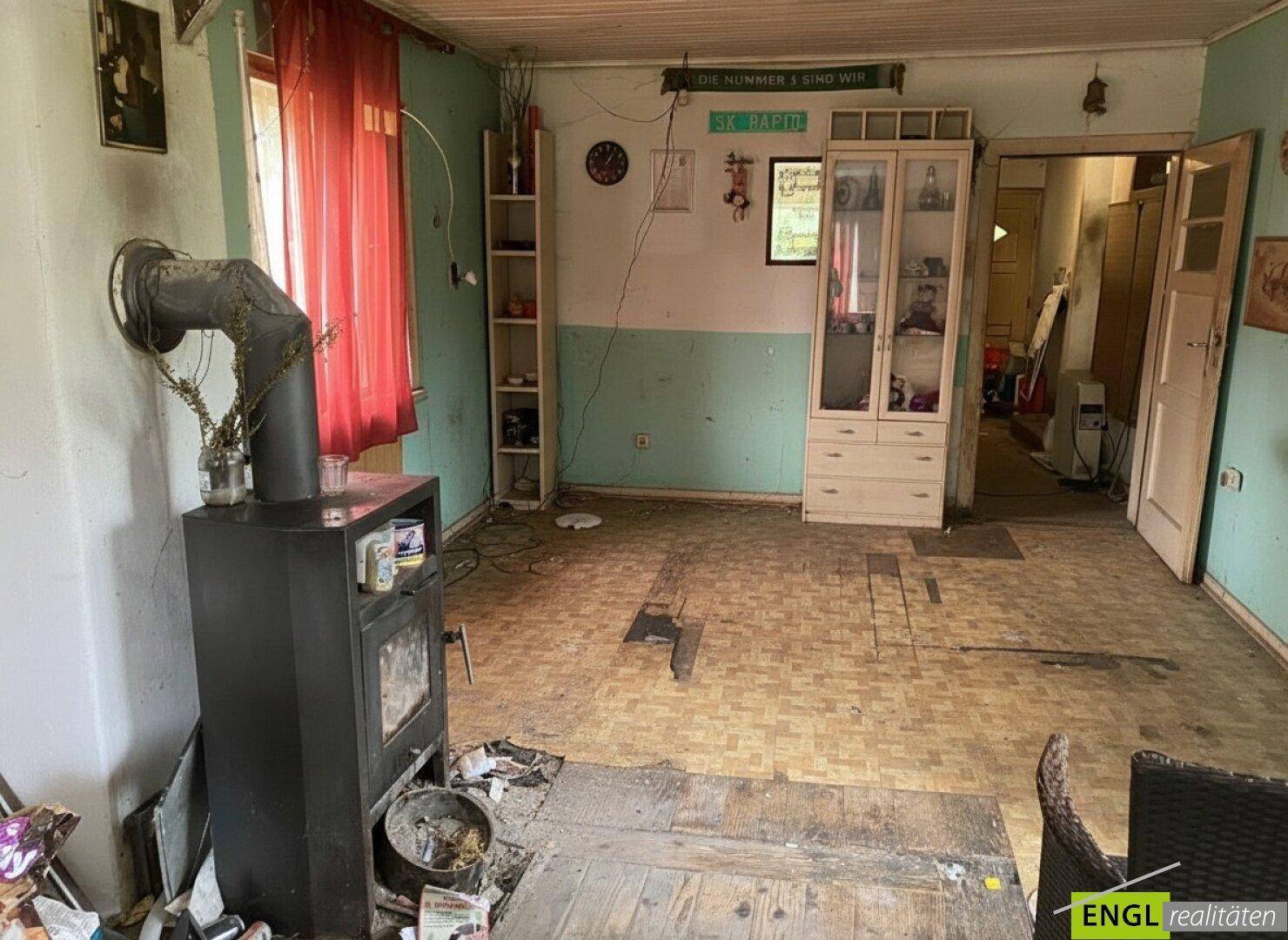 Wohnzimmer mit Holzofen, Regalen und älterem Bodenbelag, bereit für eine Renovierung.