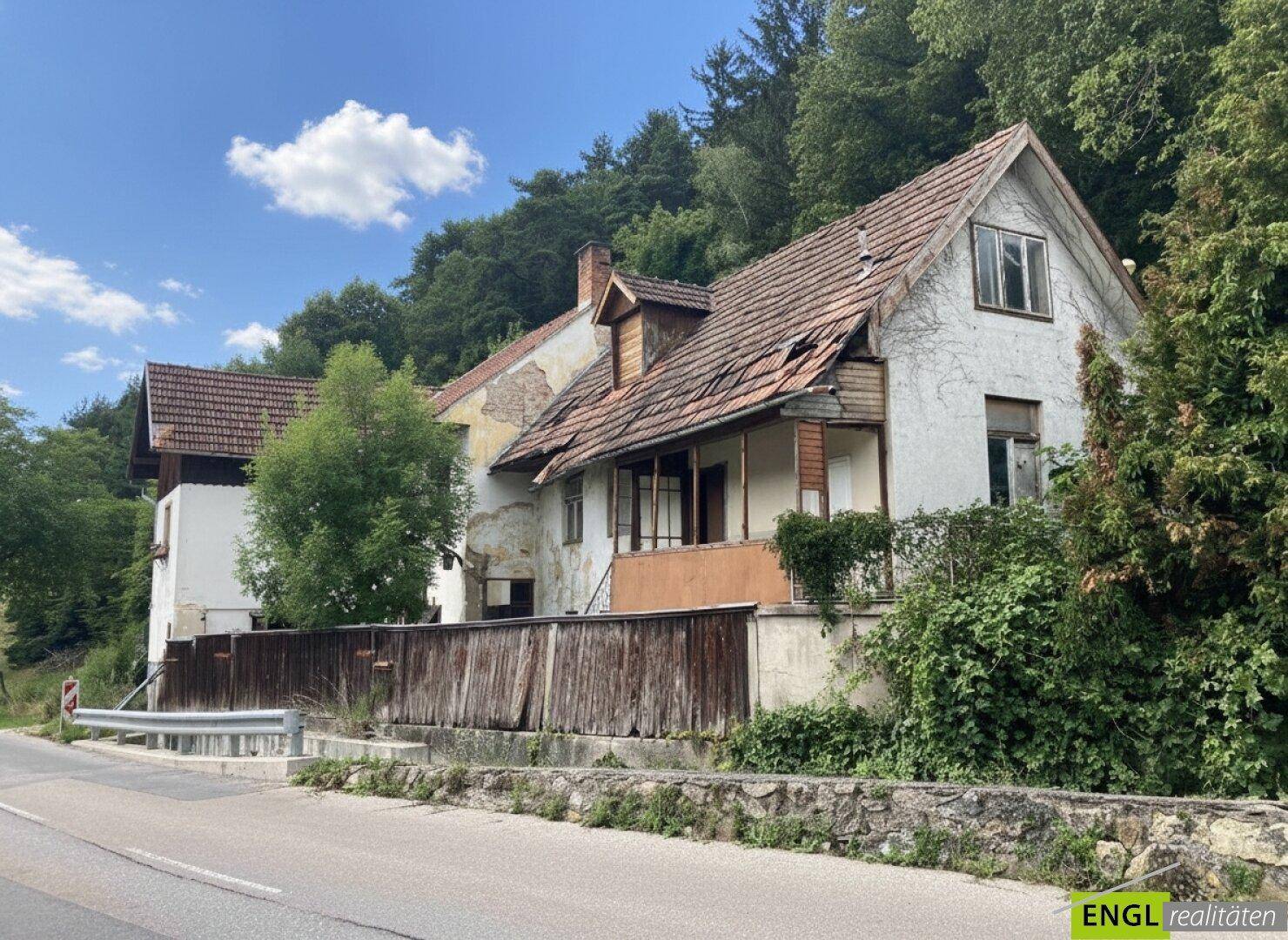 Älteres Wohnhaus mit Putzfassade, Holzzaun und Blick auf bewaldete Hügel.