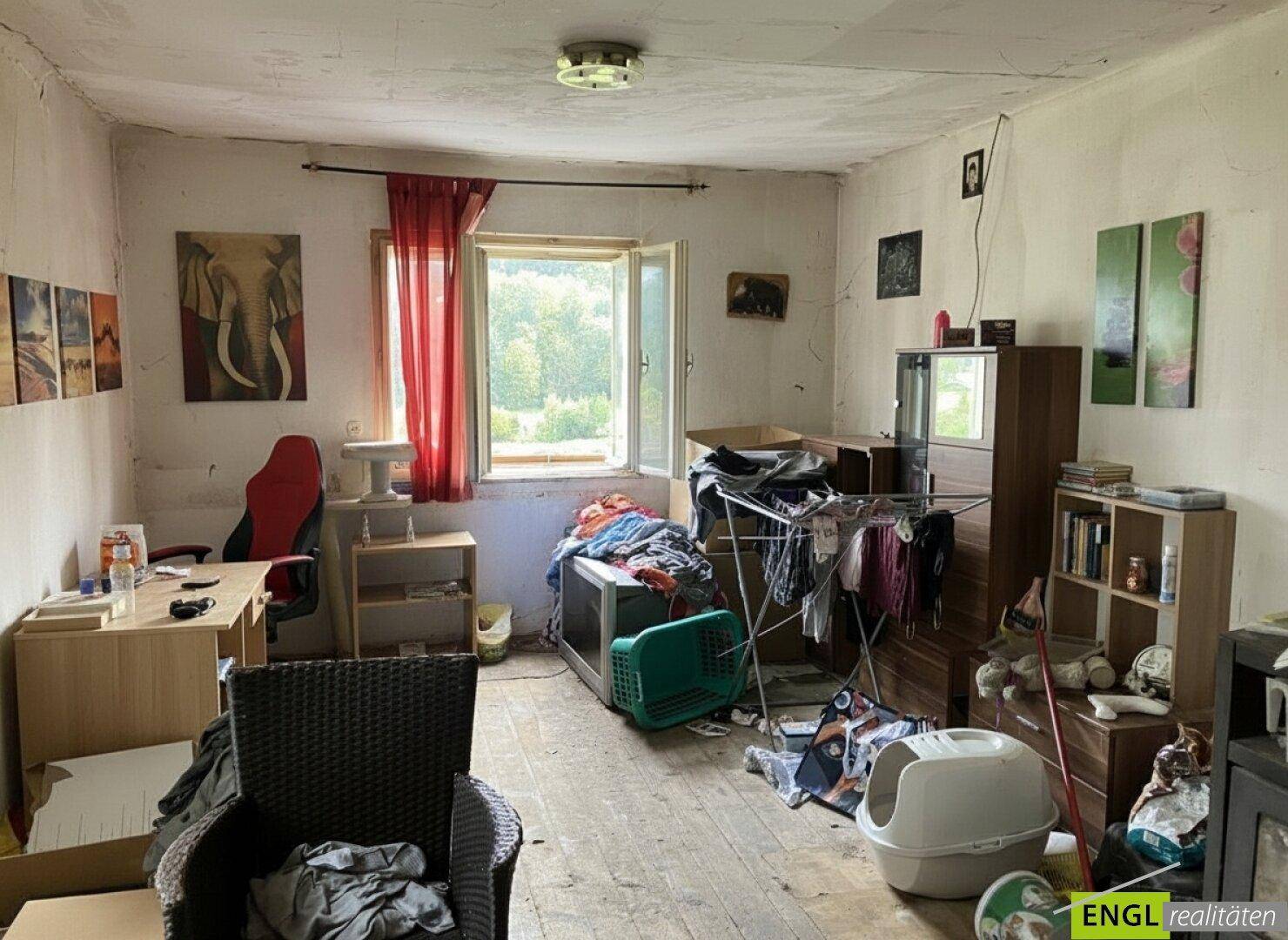 Unaufgeräumtes Schlafzimmer mit Holzdielenboden, Schreibtisch und Blick ins Grüne.