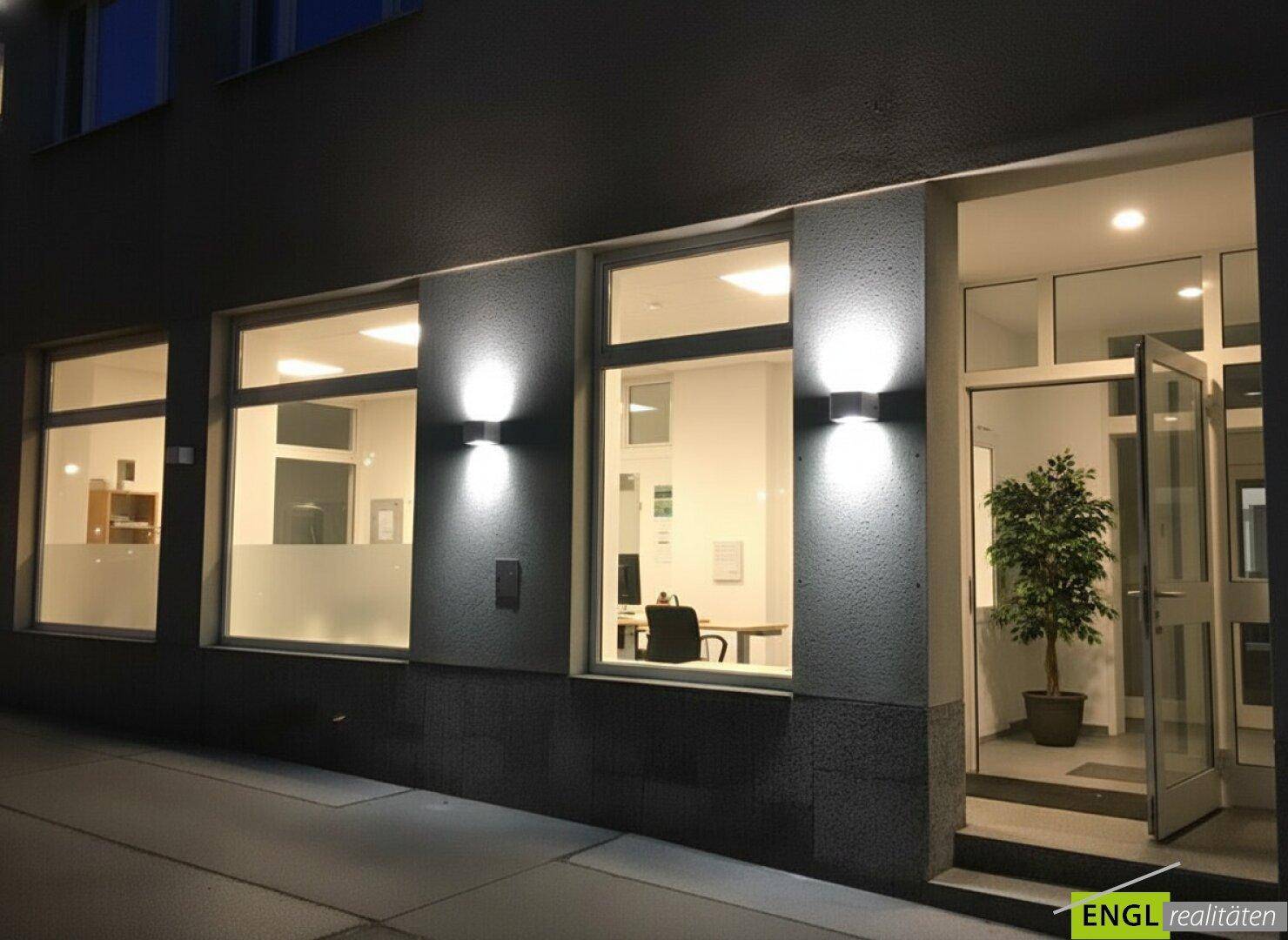 Eingangsbereich eines Geschäftsgebäudes bei Nacht, beleuchtet mit Wandlampen und großen Schaufenstern.