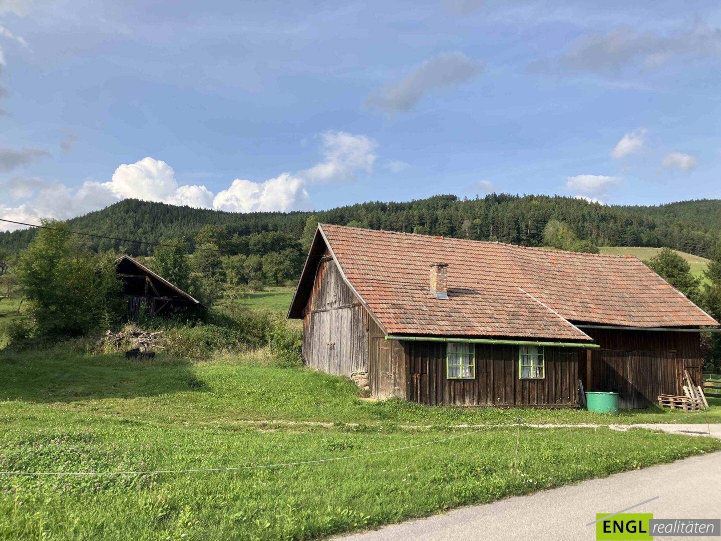 Rustikales Holzgebäude mit Ziegeldach inmitten grüner Wiesen und bewaldeter Hügel.
