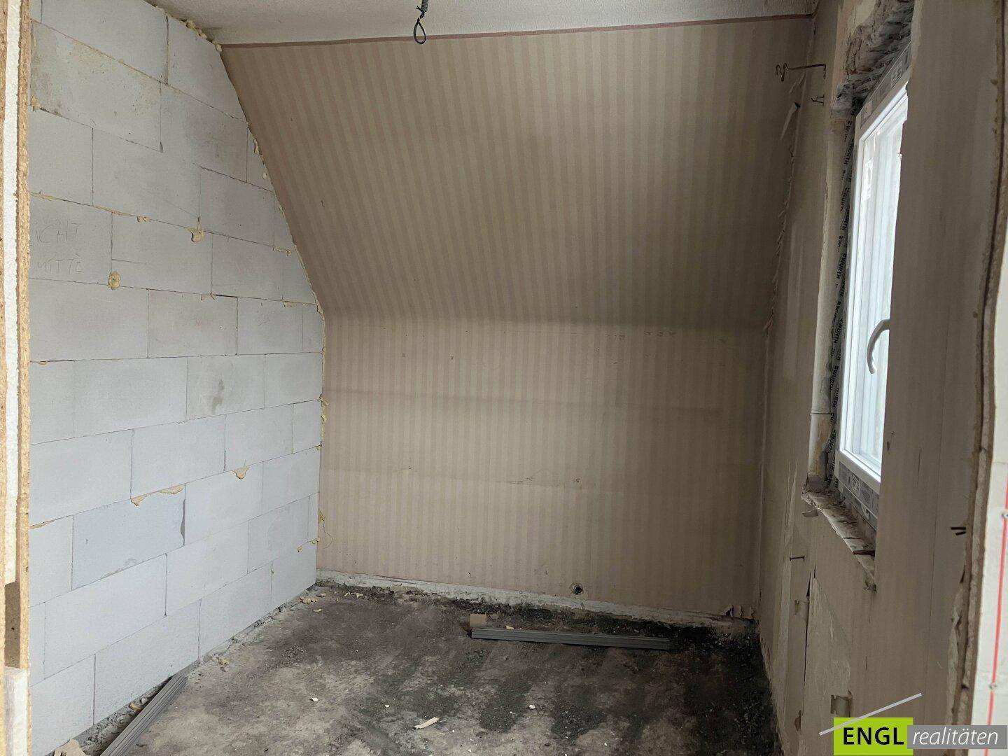 Dachgeschossraum im Rohbau mit schräger Decke und einem Fenster, bereit für den Ausbau.