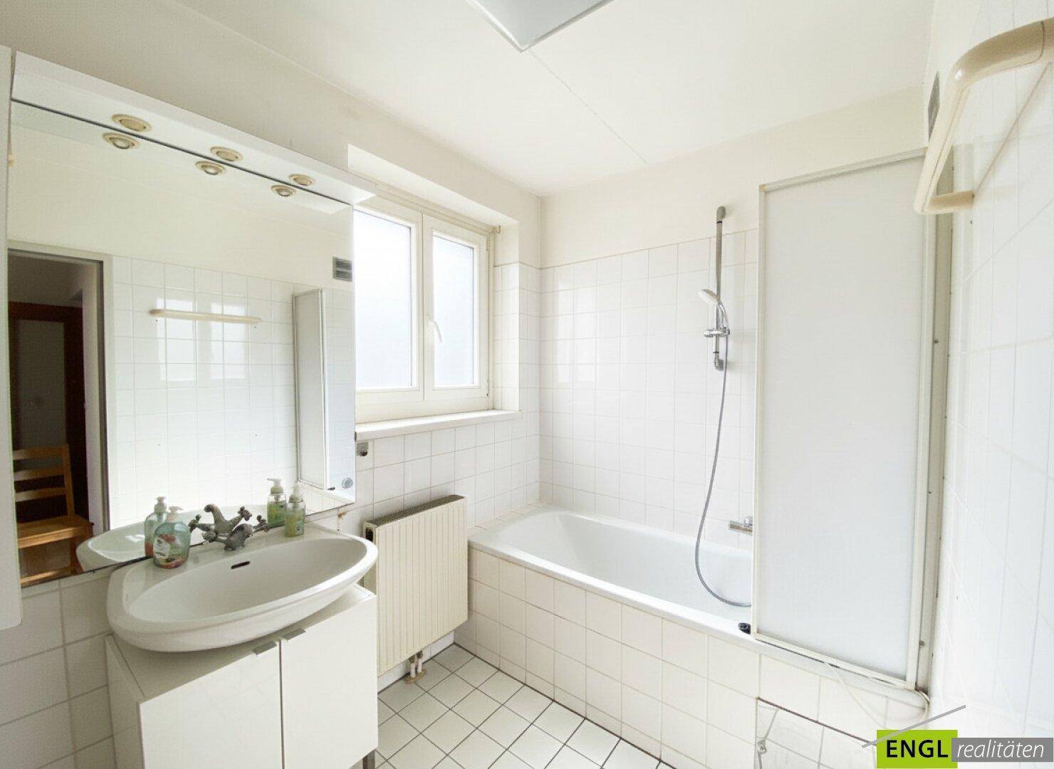 Helles Badezimmer mit Badewanne, Waschbecken, Spiegel und Fenster, komplett gefliest.