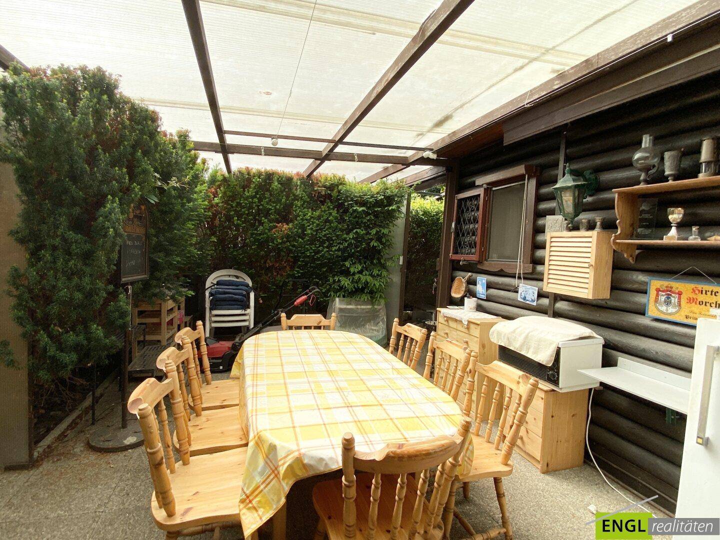 Überdachter Essbereich im Freien mit Holztisch und Stühlen, angrenzend an eine rustikale Holzhütte.