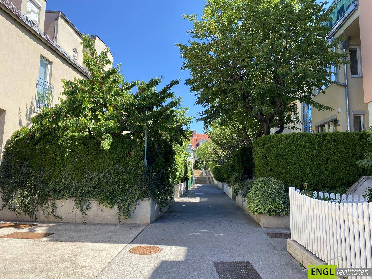 Ruhiger Fußweg zwischen gepflegten Hecken und Bäumen, der zu den Wohnhäusern führt.
