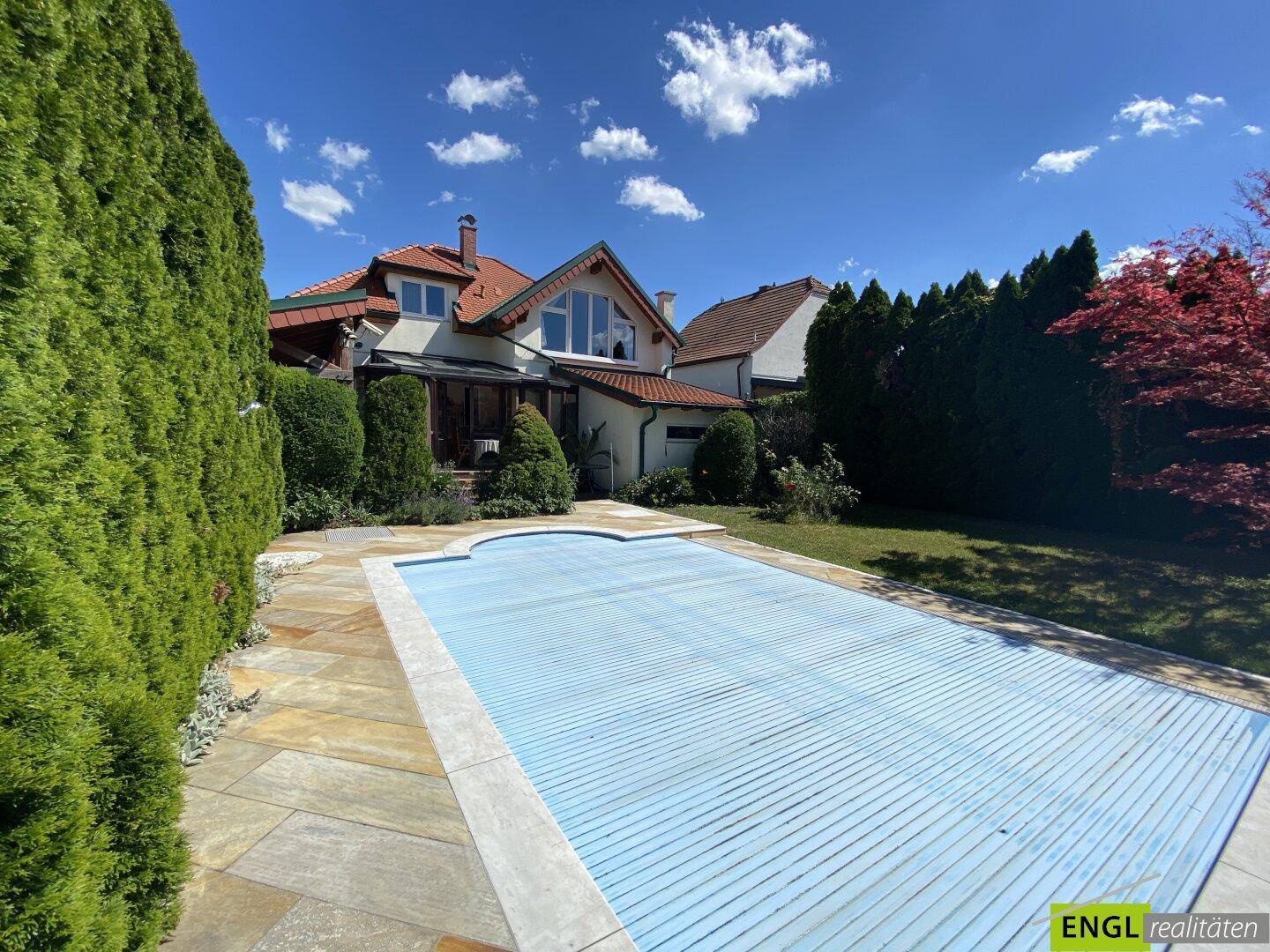Hausfassade mit großem Garten und abgedecktem Swimmingpool an einem sonnigen Tag.