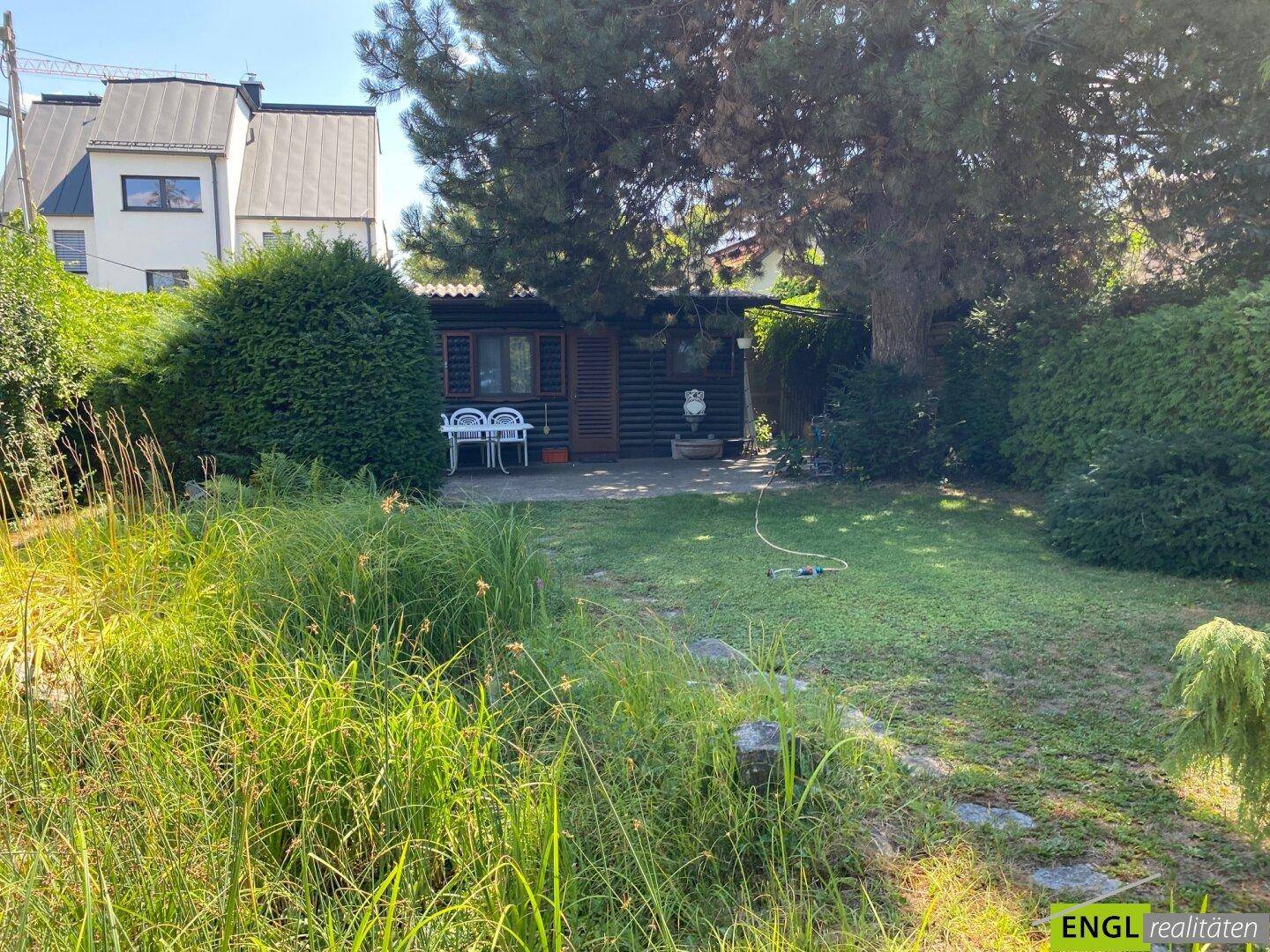 Gepflegter Garten mit Rasenfläche, schattenspendenden Bäumen und einem gemütlichen Gartenhaus mit Sitzgelegenheit.