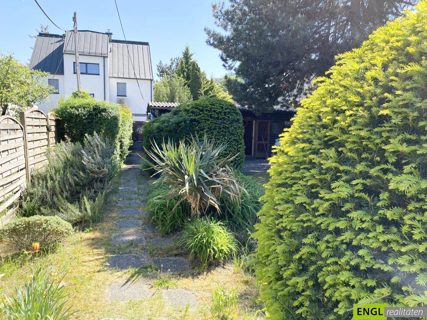 Gartenweg durch eine vielfältige Bepflanzung, der zu einem Wohnhaus im Hintergrund führt.