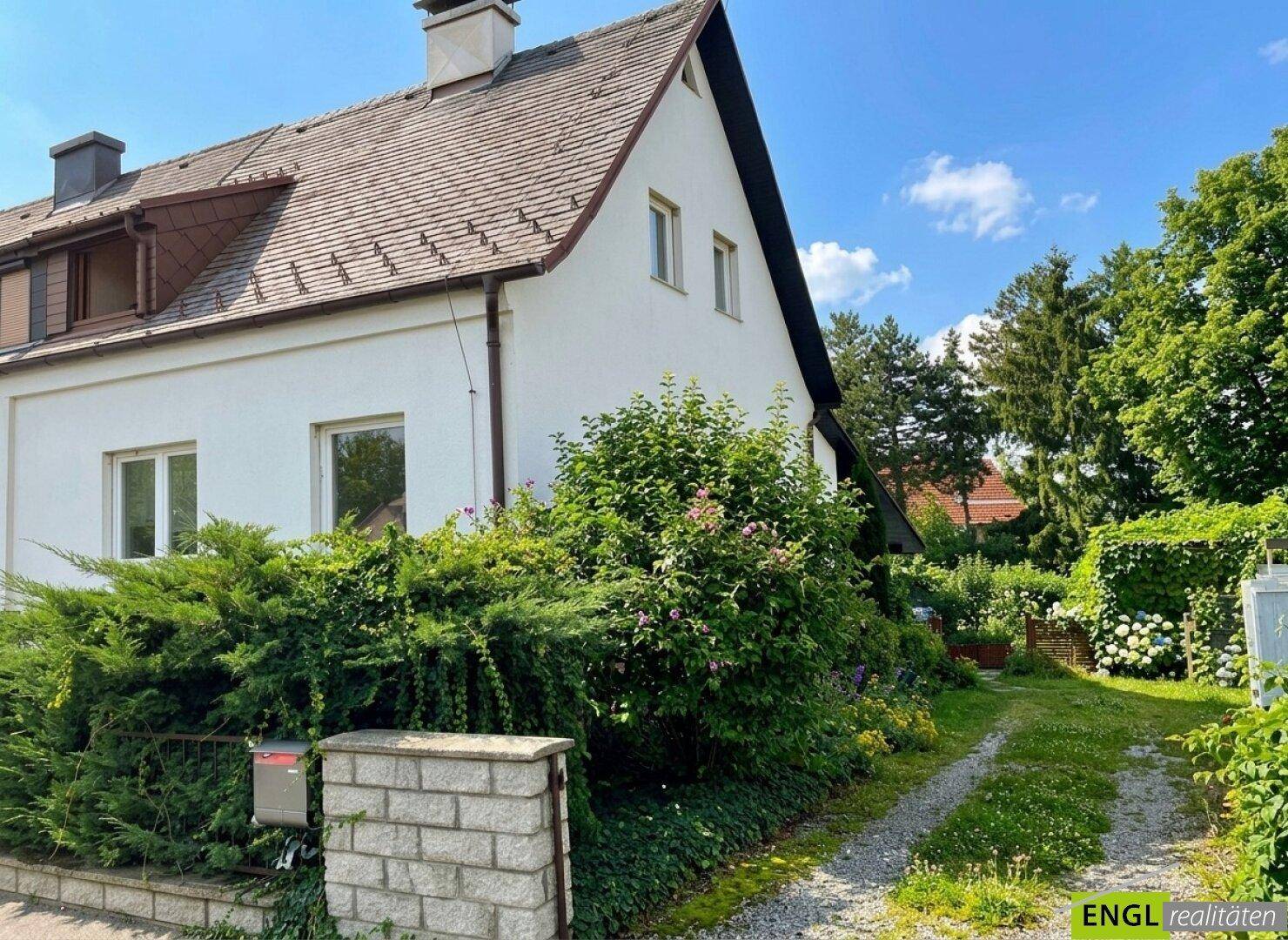 Seitenansicht des Hauses mit Kiesauffahrt und üppigem Grün im Vorgarten bei Sonnenschein.