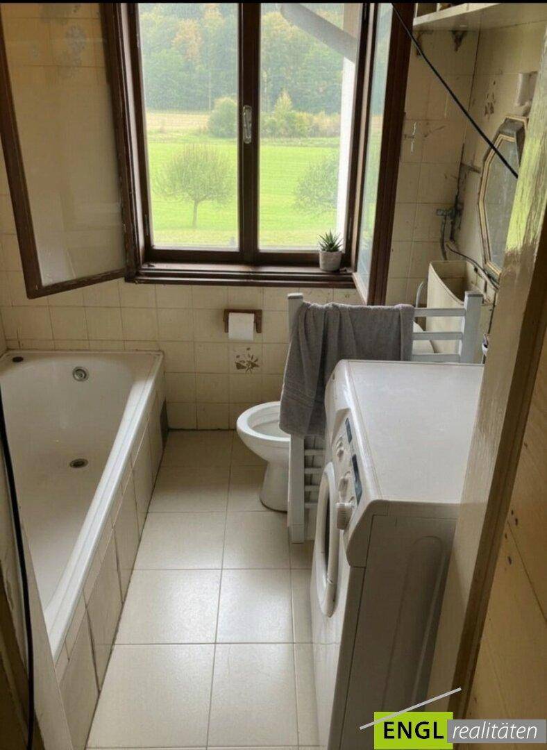 Badezimmer mit Badewanne, Toilette, Waschmaschine und Fenster mit Blick ins Grüne.