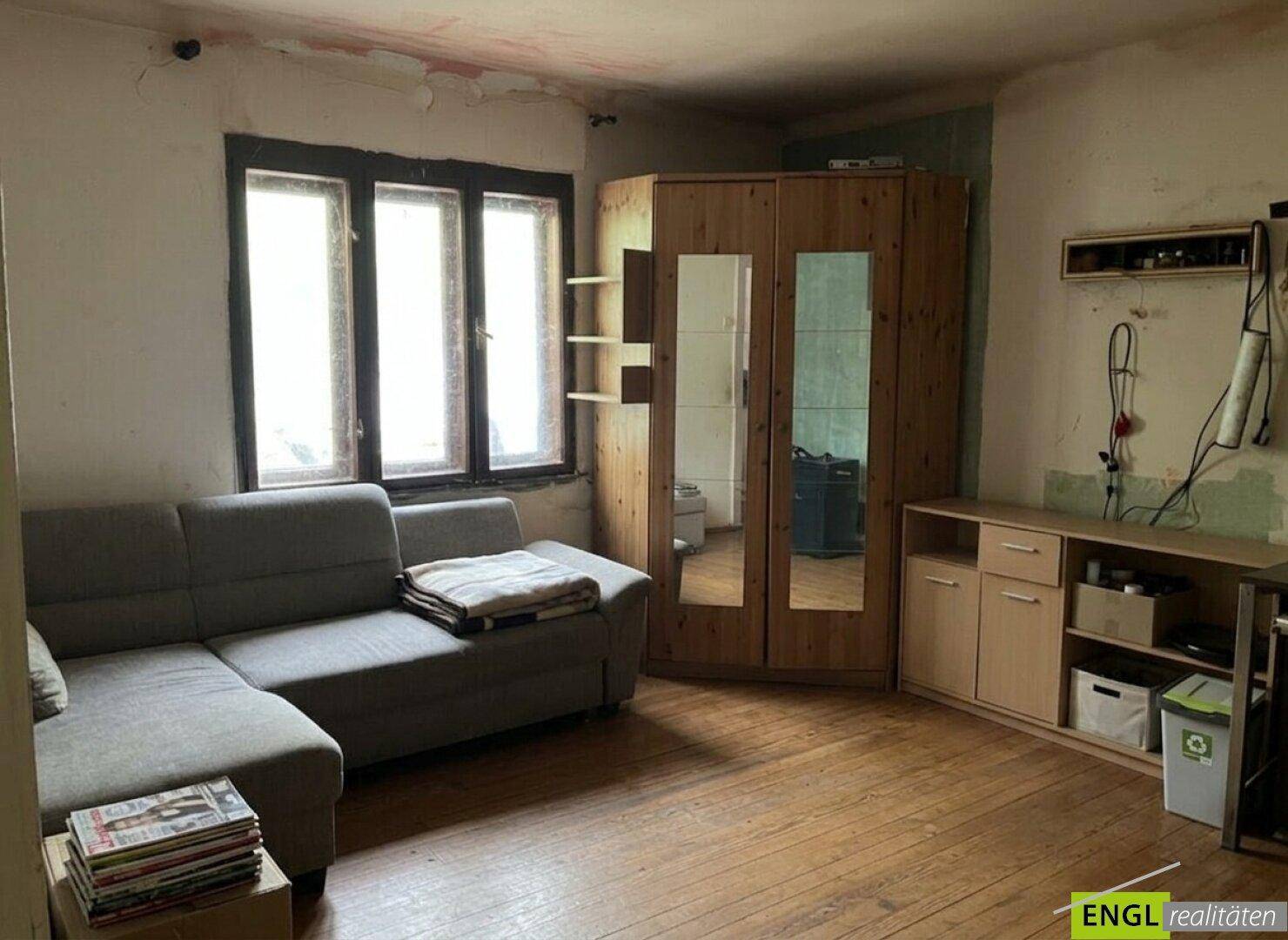 Einfaches Wohnzimmer mit Sofa, Holzschrank und Fenster, das auf eine Renovierung wartet.