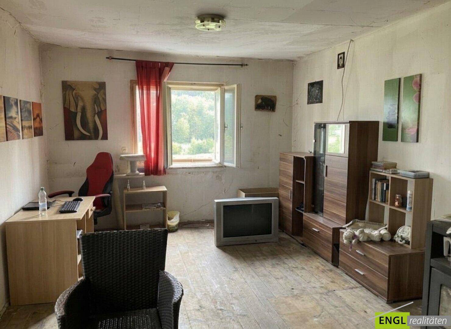 Abgenutztes Wohnzimmer mit alten Möbeln, einem Schreibtisch und Blick ins Grüne.