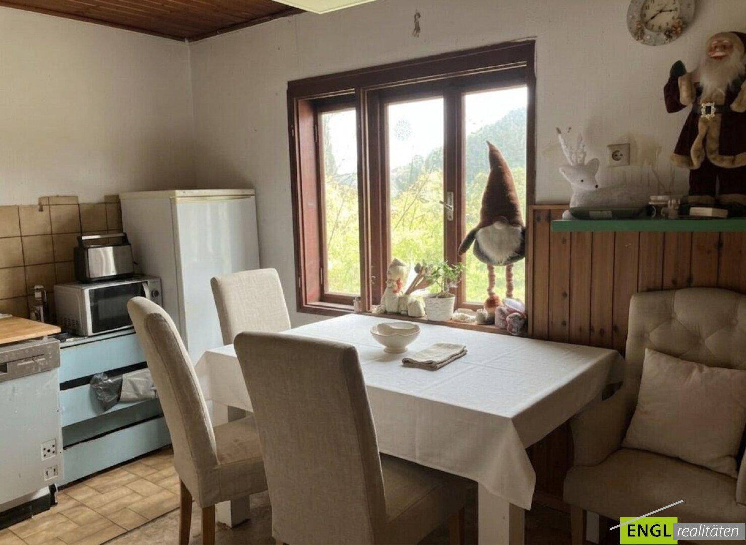 Einfacher Essbereich mit Tisch, Stühlen, Kühlschrank und Fensterblick auf die Natur.