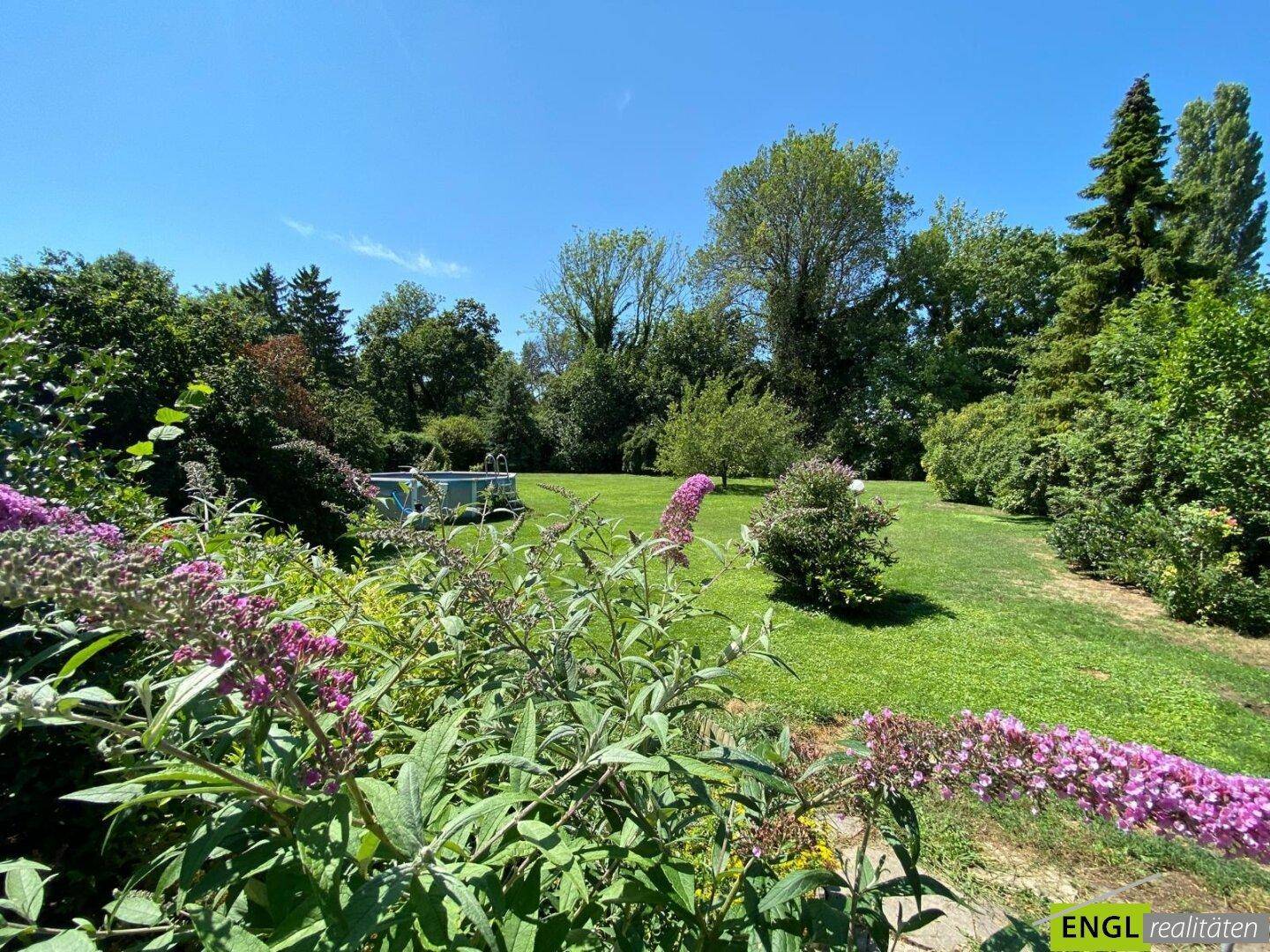 Weitläufiger Garten mit üppiger Vegetation, Rasenflächen und einem kleinen Pool im Hintergrund.