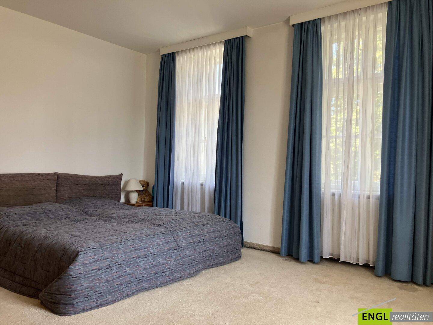 Geräumiges Schlafzimmer mit großem Bett, Teppichboden und hohen Fenstern mit blauen Vorhängen.