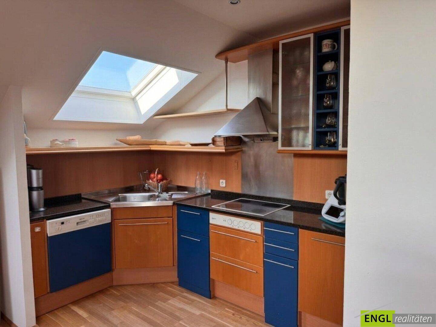 Funktionale Küche mit blauen und holzfarbenen Schränken, dunkler Arbeitsplatte und Dachfenster.