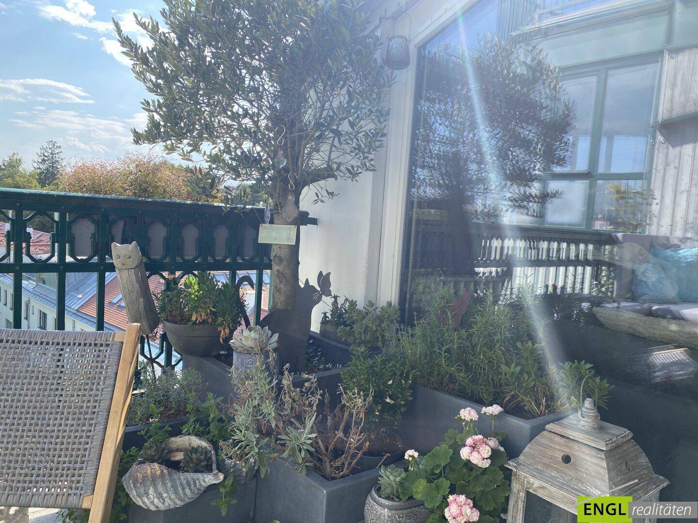 Sonniger Balkon mit üppiger Bepflanzung, einem Olivenbaum und Blick auf die Umgebung.