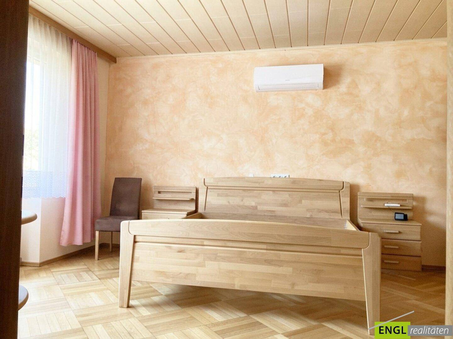 Helles Schlafzimmer mit einem Holzdoppelbett, Nachttischen und einer Klimaanlage an der Wand.