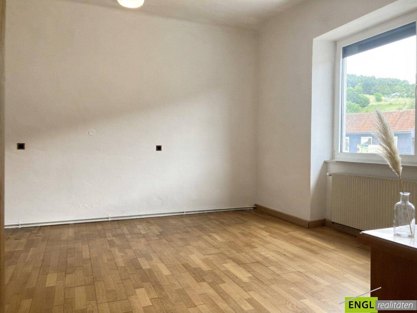 Leerer Raum mit Parkettboden und einem großen Fenster, das einen Blick ins Grüne bietet.