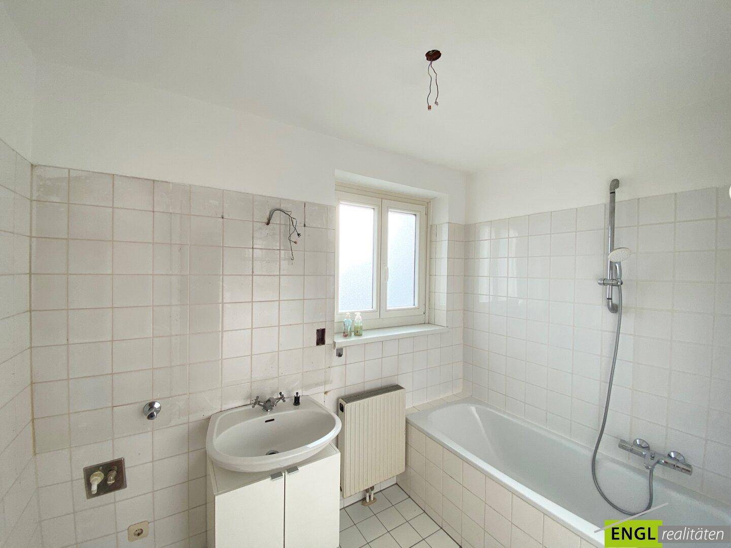 Helles Badezimmer mit weißen Fliesen, Badewanne und einem Fenster für Tageslicht.