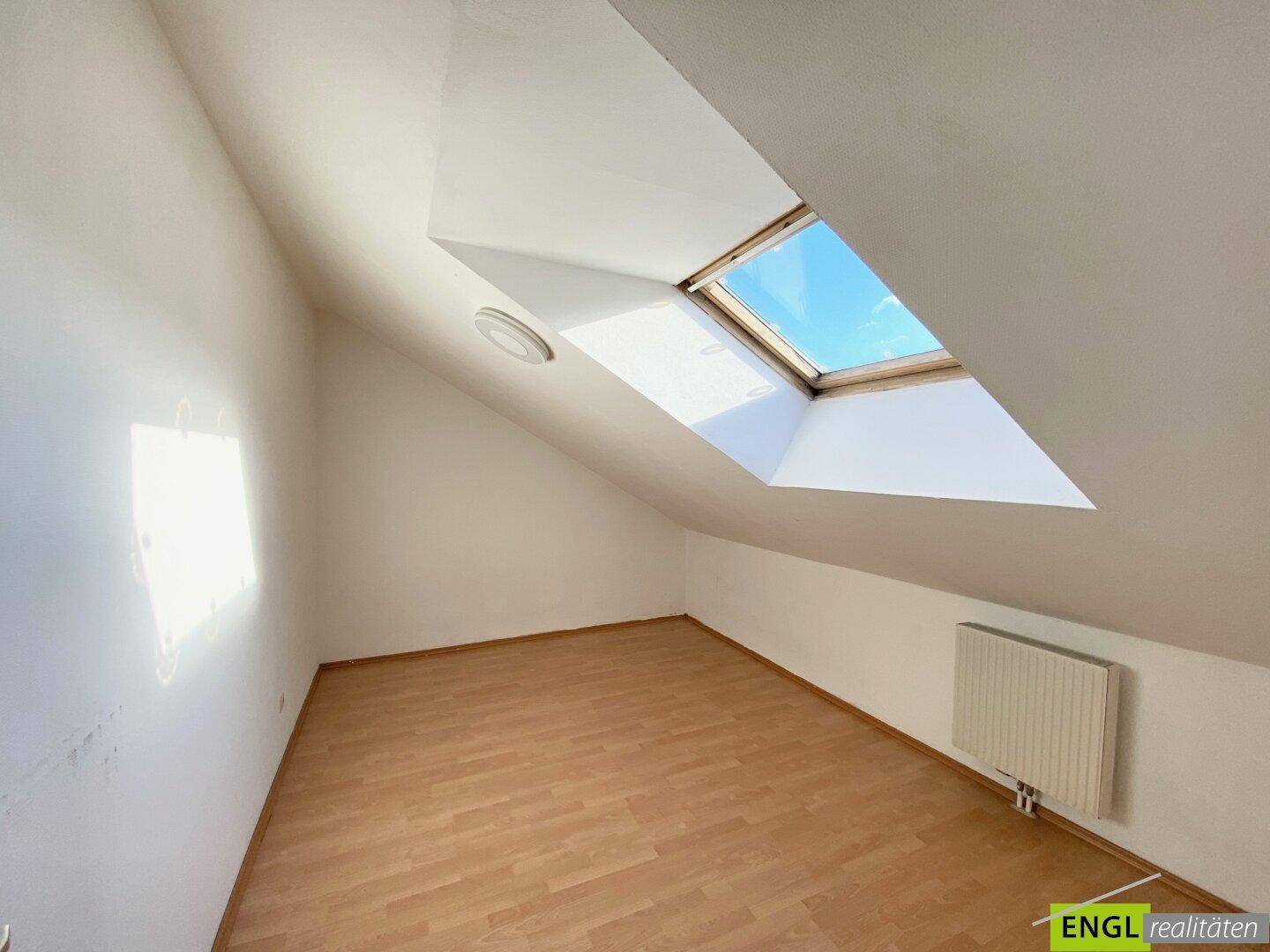 Leeres Dachzimmer mit schrägen Wänden, Holzboden und einem großen Dachfenster.