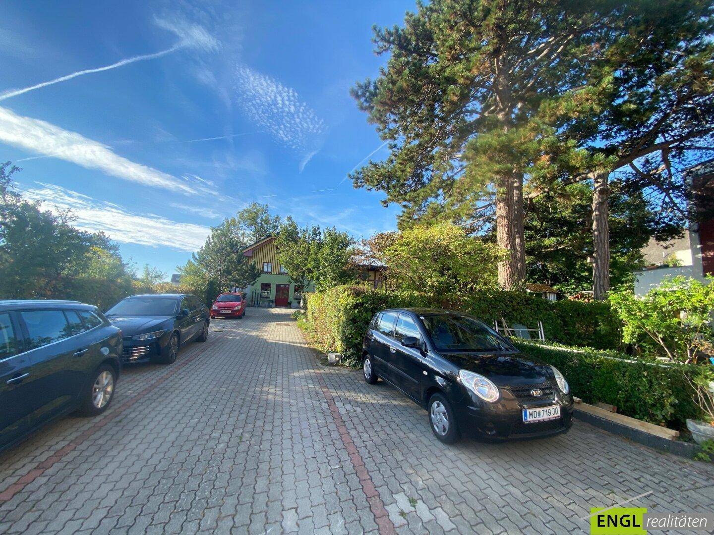 Außenparkplätze auf einem gepflasterten Hof, umgeben von Hecken und Bäumen.