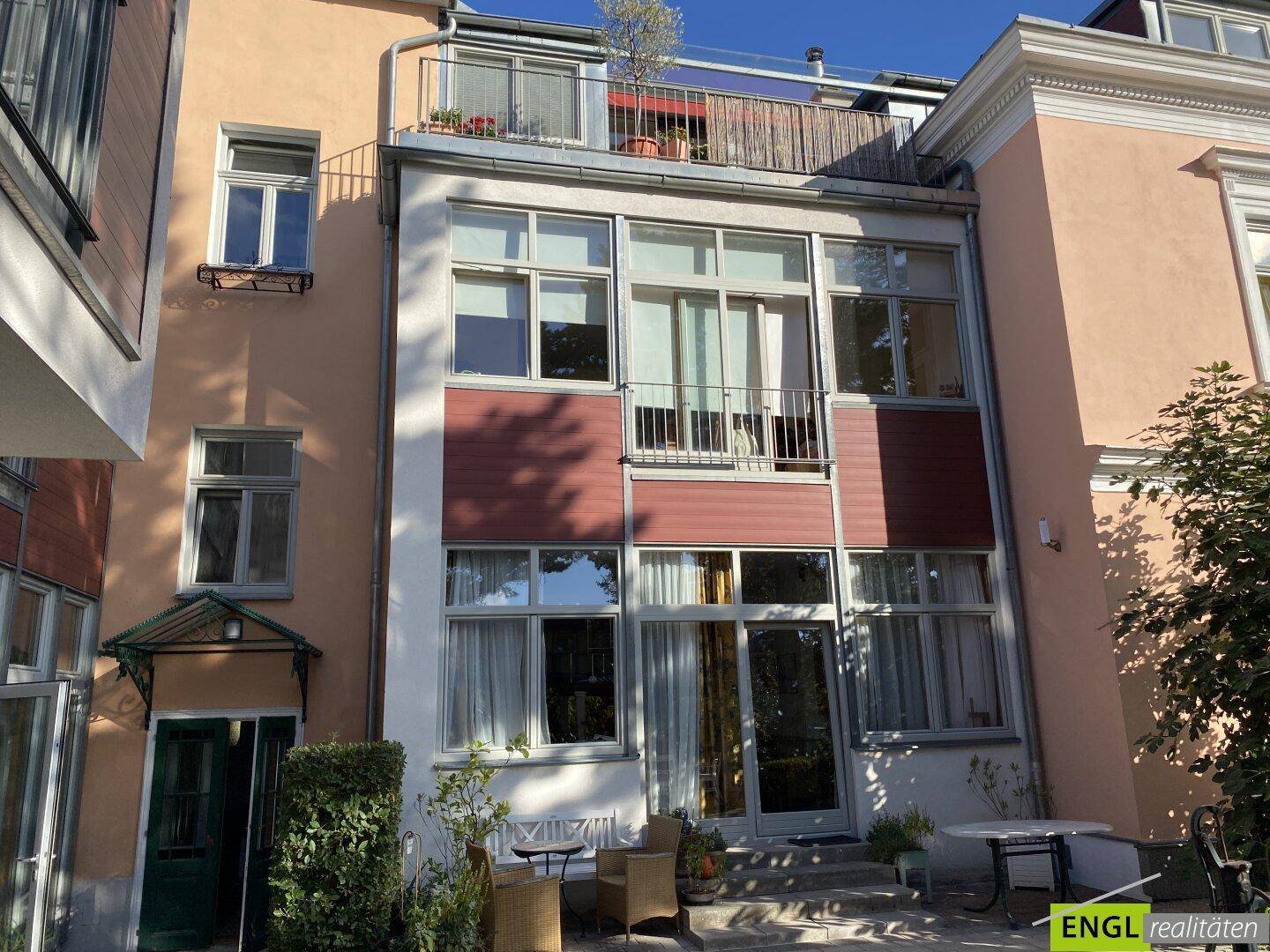 Rückseite des Gebäudes mit mehreren Fenstern, einem Balkon und einer kleinen Terrasse im Erdgeschoss.