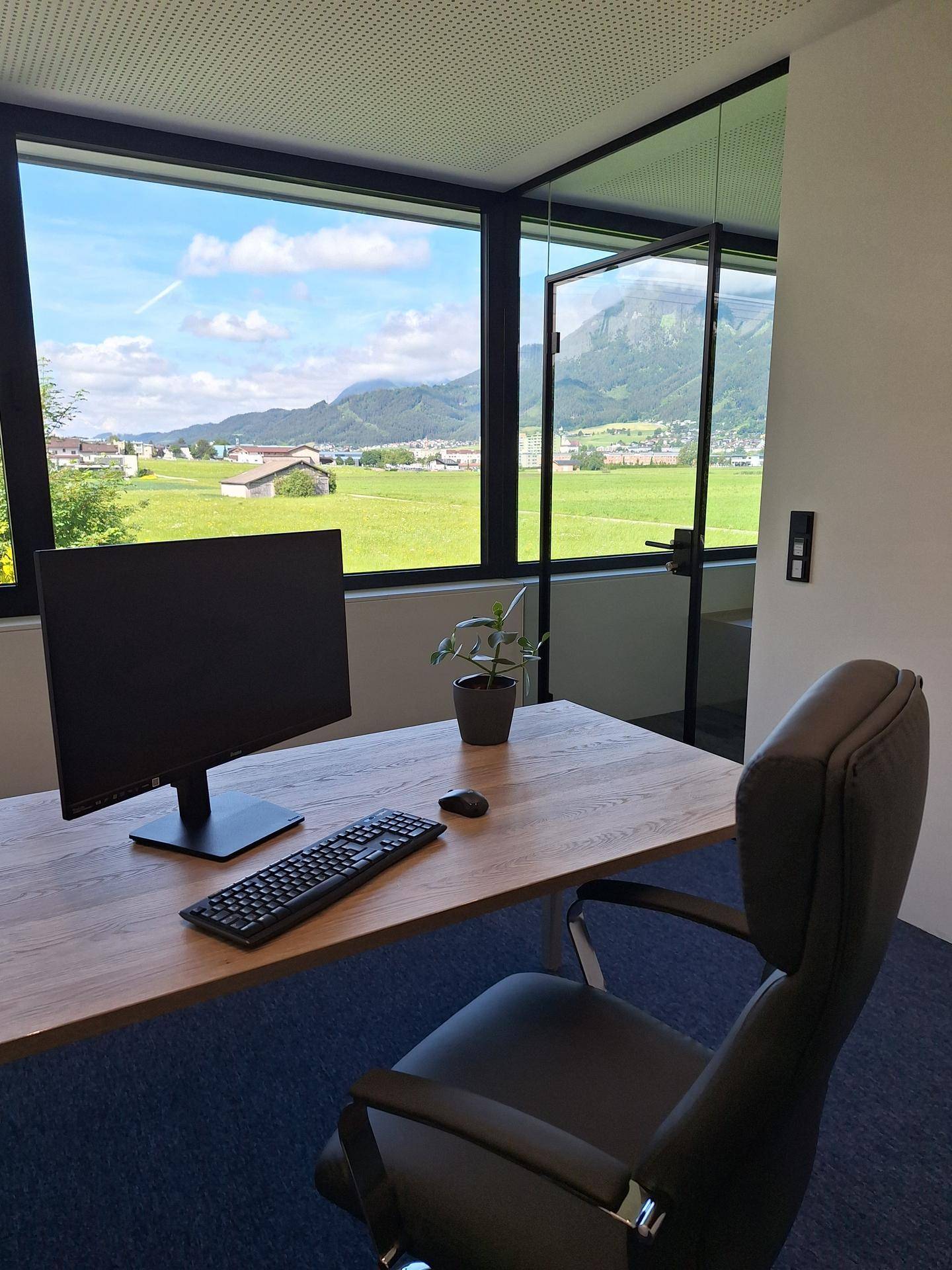 Heller Büroraum mit großem Fenster, das einen weiten Blick auf grüne Felder und Berge bietet.