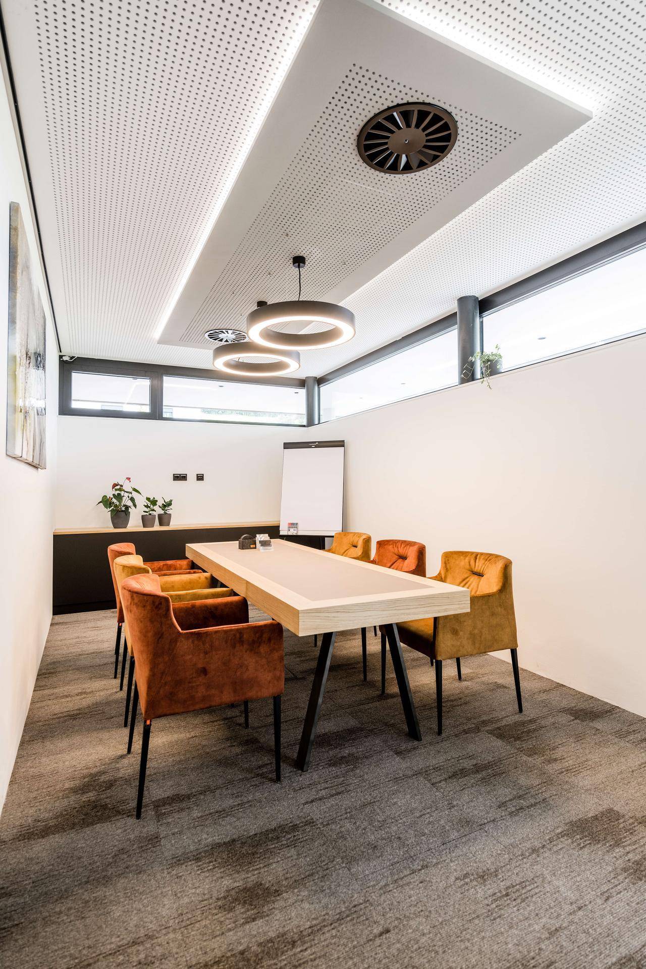 Moderner Konferenzraum mit langem Holztisch, orangefarbenen Samtstühlen und perforierter Decke.
