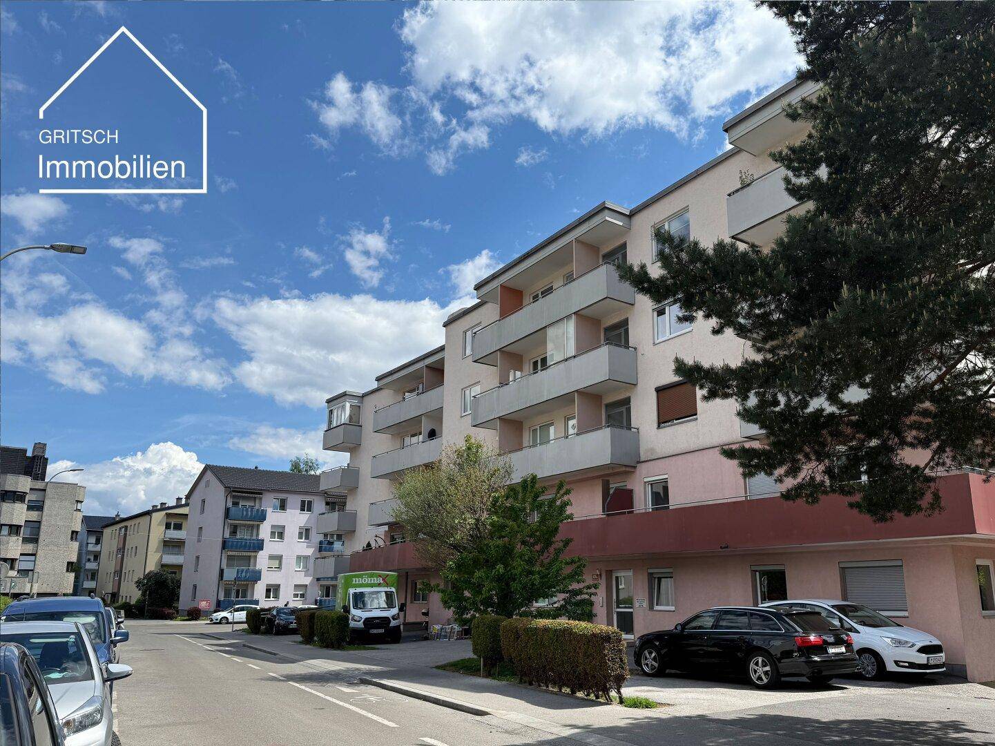 Ansicht eines Wohngebäudes mit Balkonen und Bäumen an der Straßenseite unter einem bewölkten Himmel.